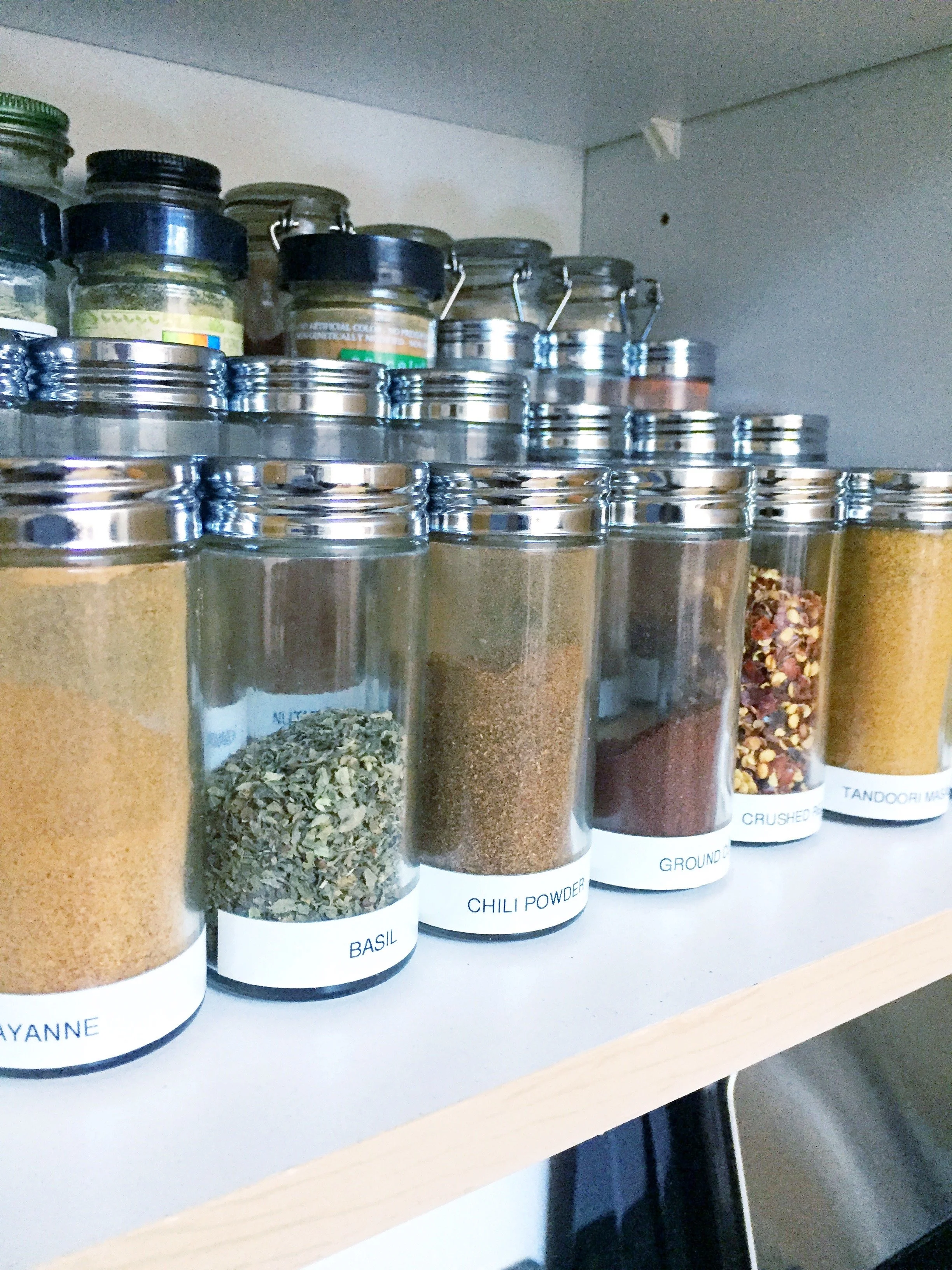 Kitchen cabinet with jars of spices labeled basil, chili powder, ground cumin, crushed red pepper, and tandoori masala.