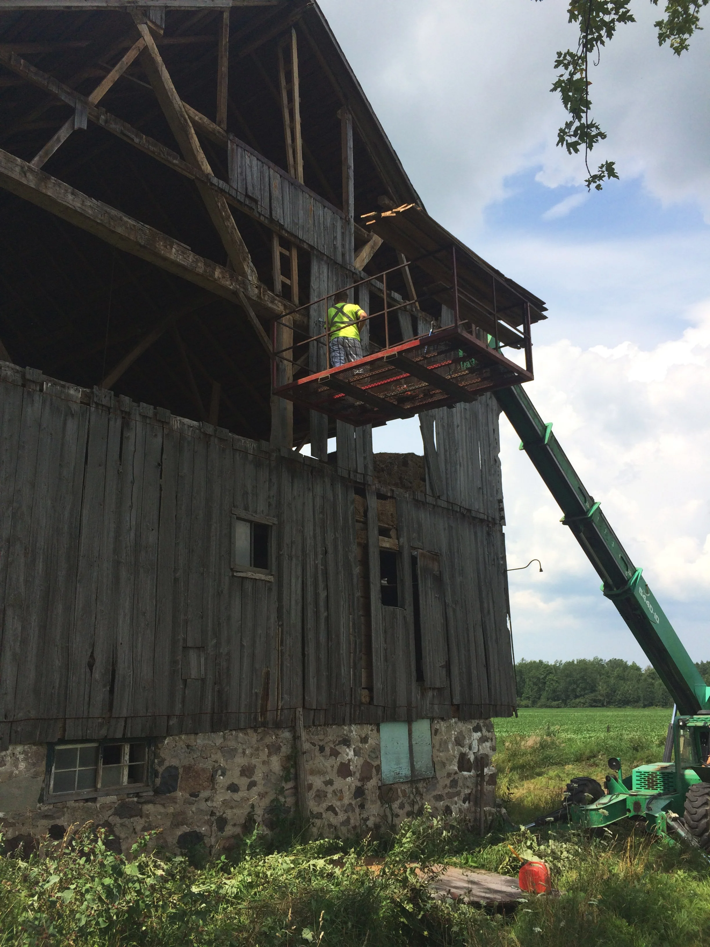A worker in a yellow safety vest on a raised work platform cleaning or inspecting the upper part of a weathered barn with wooden siding and stone foundation, with a telescoping boom lift in a rural outdoor setting under a partly cloudy sky.