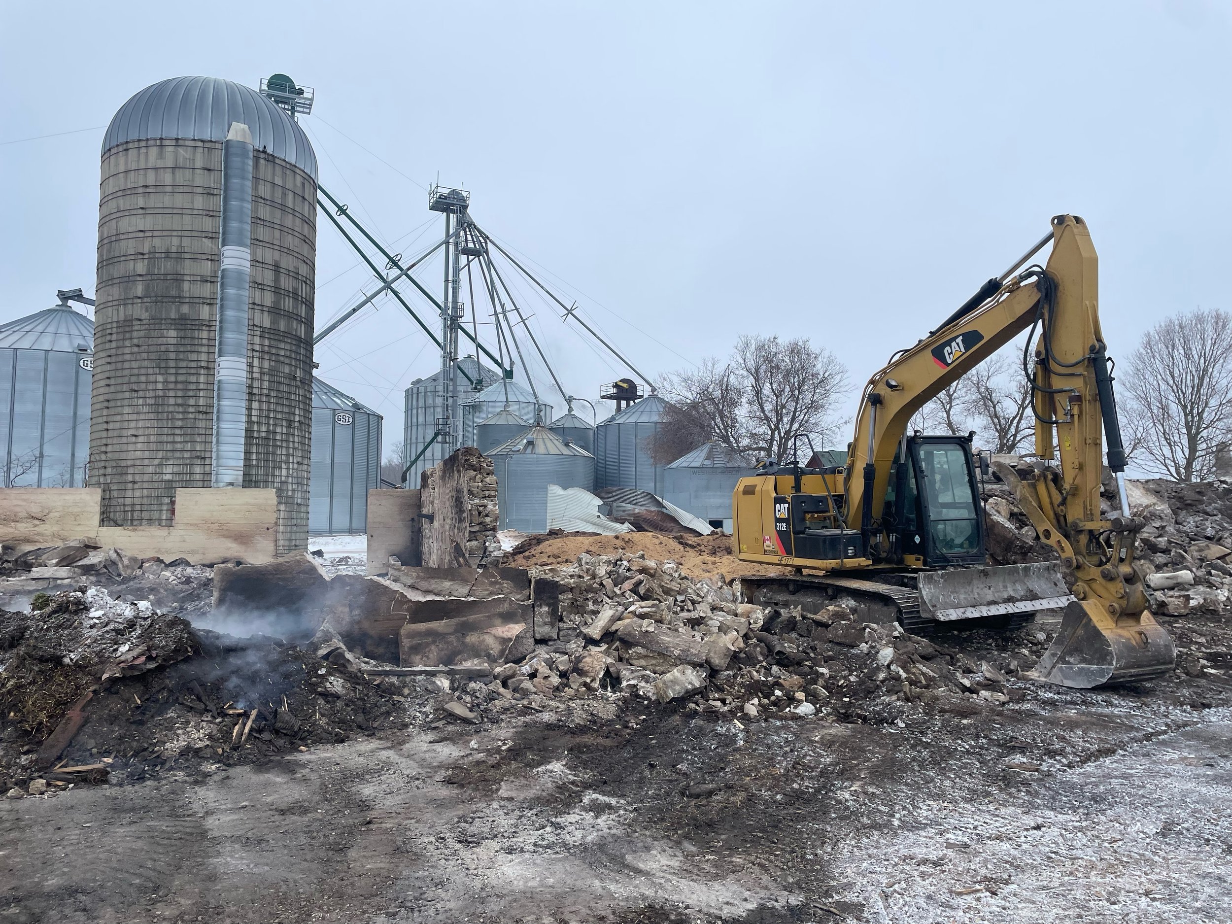 Construction site with a yellow excavator working on the ground, and multiple grain silos in the background under an overcast sky.