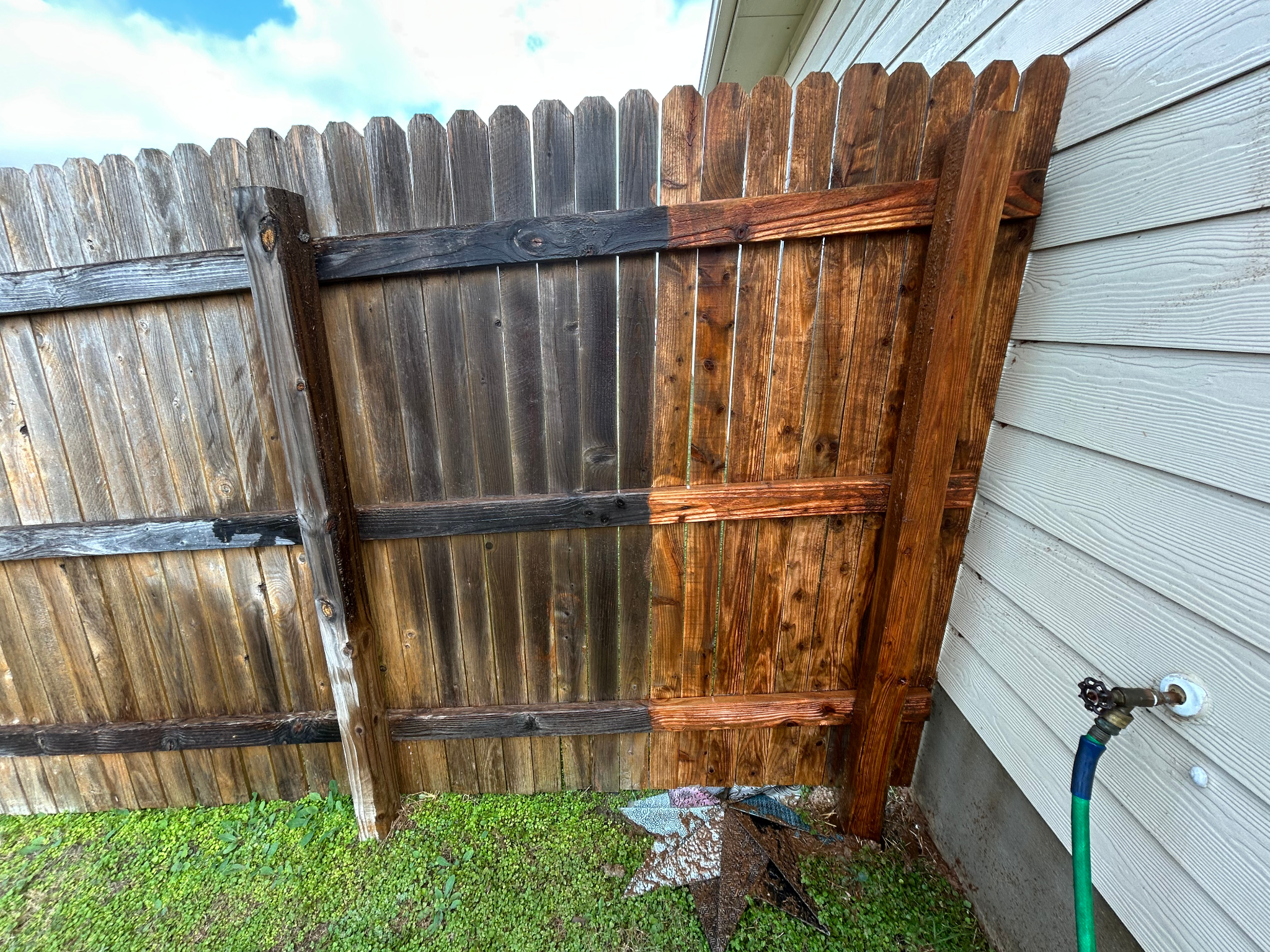 A newly built wooden fence gate attached to a house exterior with siding, next to a faucet with a garden hose, and a small patch of grass on the ground.