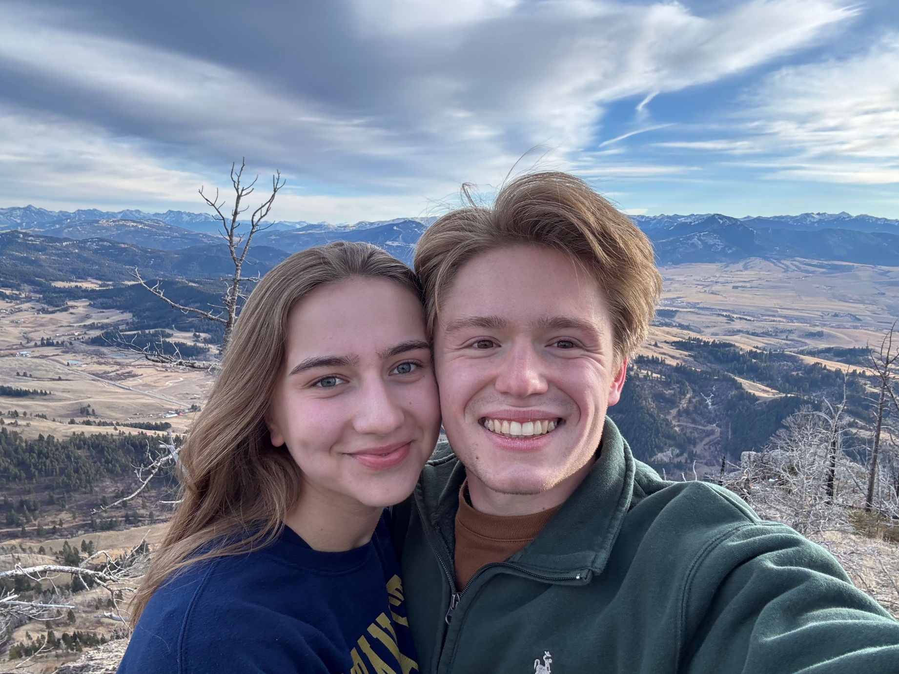 A smiling young man and woman taking a selfie against a mountain landscape with snow-capped peaks, fields, and trees in the background.