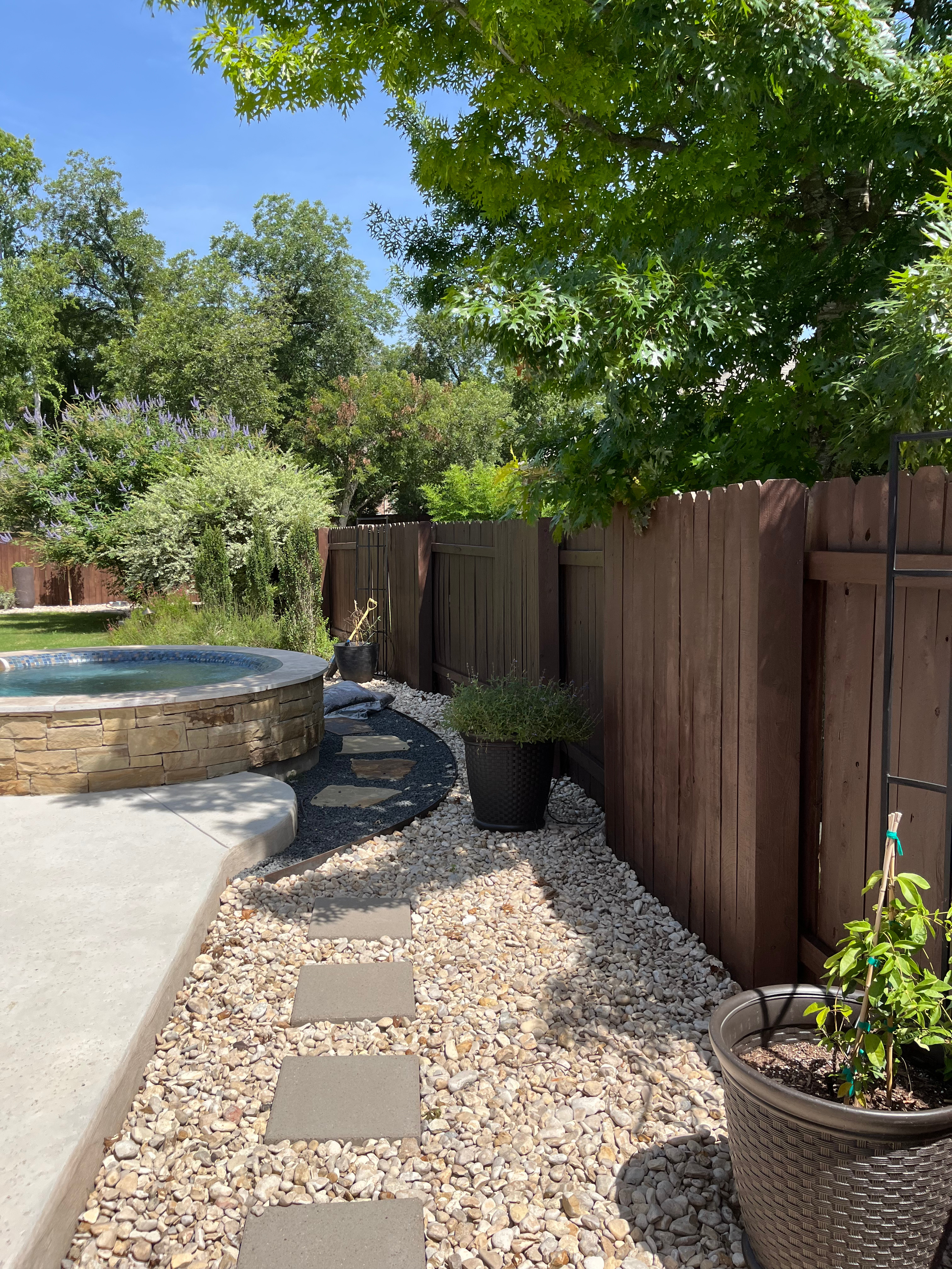 A backyard with a stone-lined hot tub, stepping stones, potted plants, a wooden fence, and a large leafy tree providing shade.