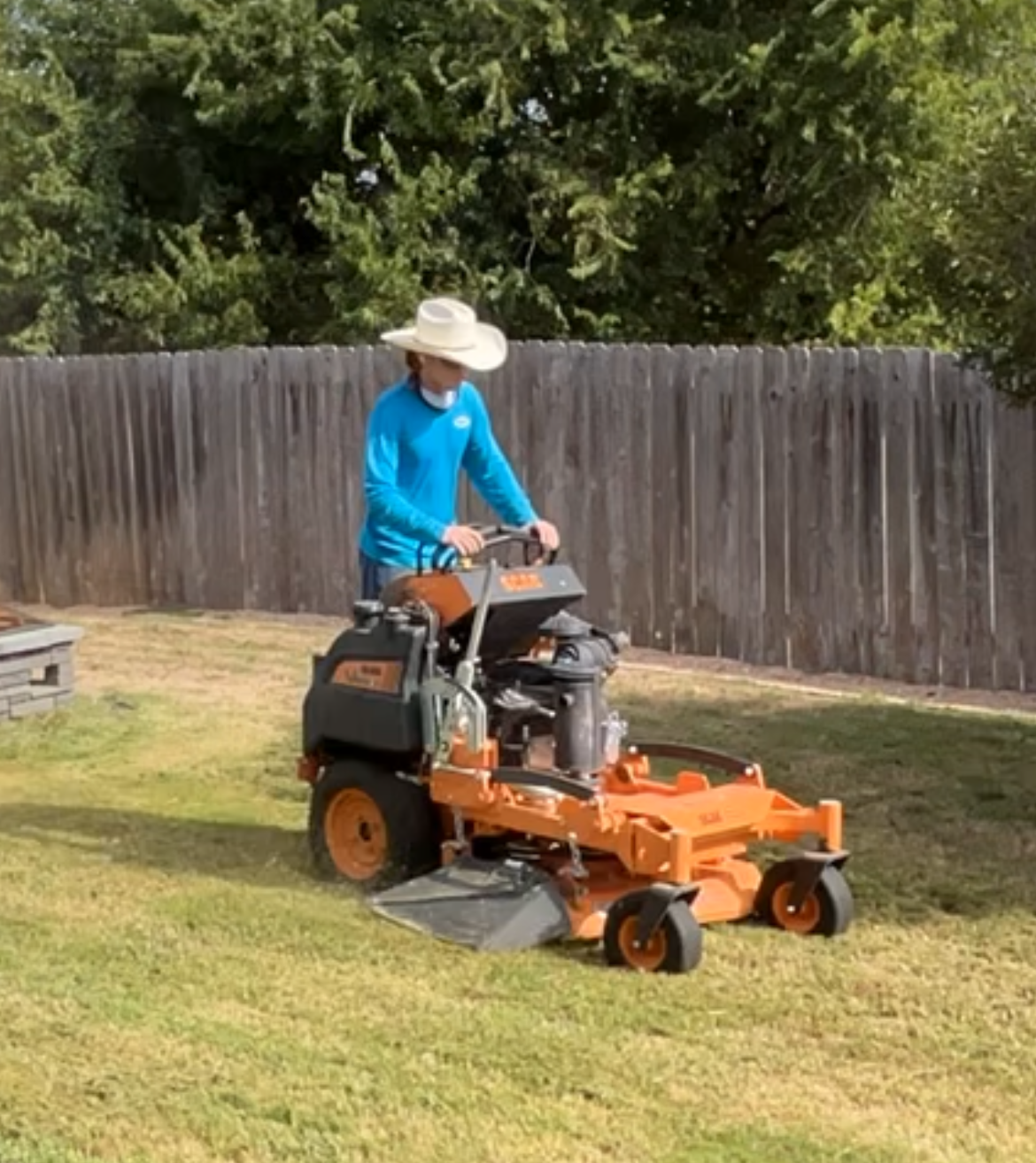 Person wearing a large sun hat and blue long-sleeve shirt riding an orange commercial zero-turn lawn mower on a grassy yard enclosed by a wooden fence and trees.