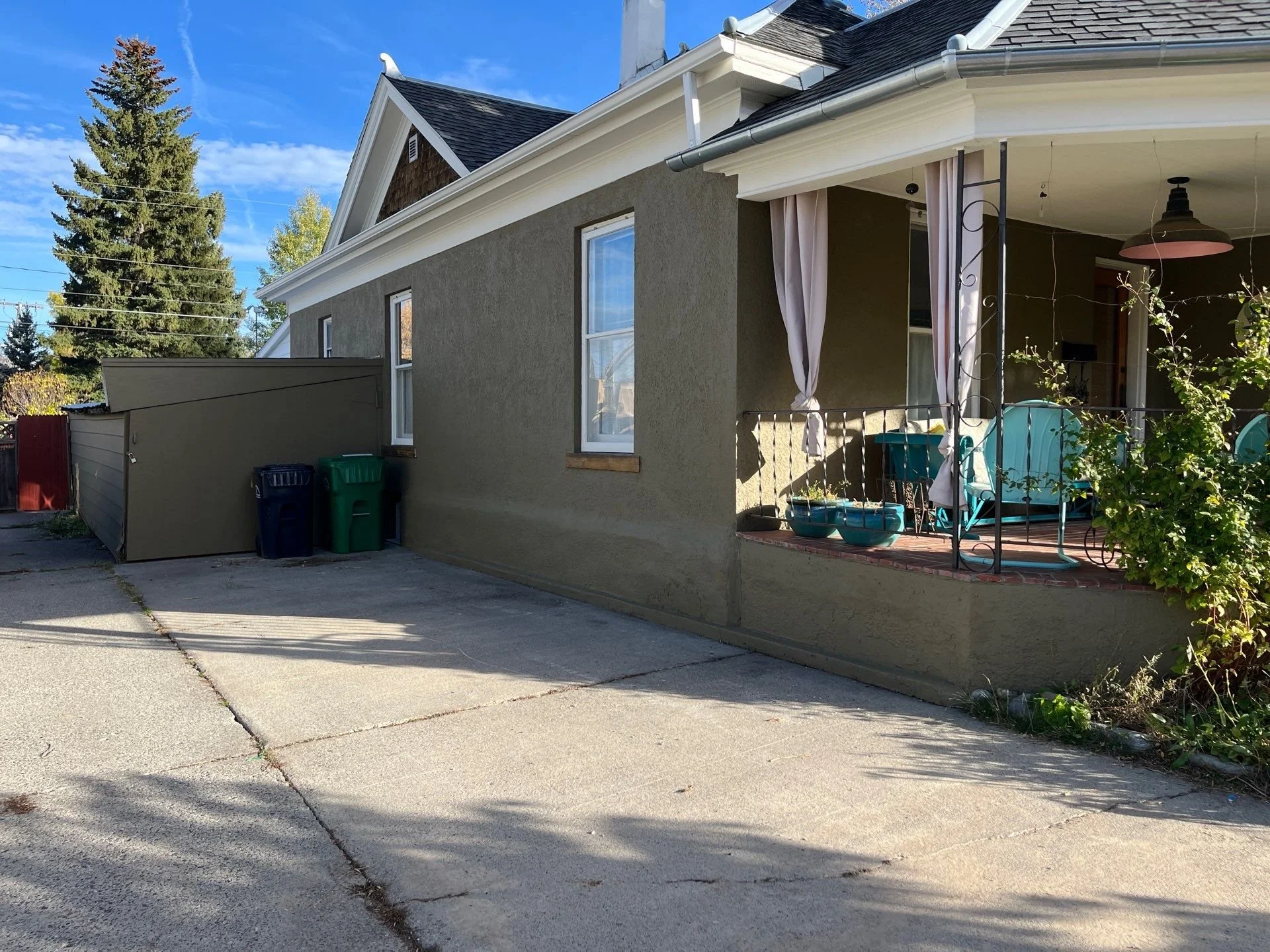 A house with a small front porch, two turquoise chairs, parked plants, and hanging curtains. The house has a grayish exterior wall, two windows, and a sloped roof with white trims. A concrete driveway extends in front of the house, with some shadows cast on it. There are tall trees and a blue sky in the background.