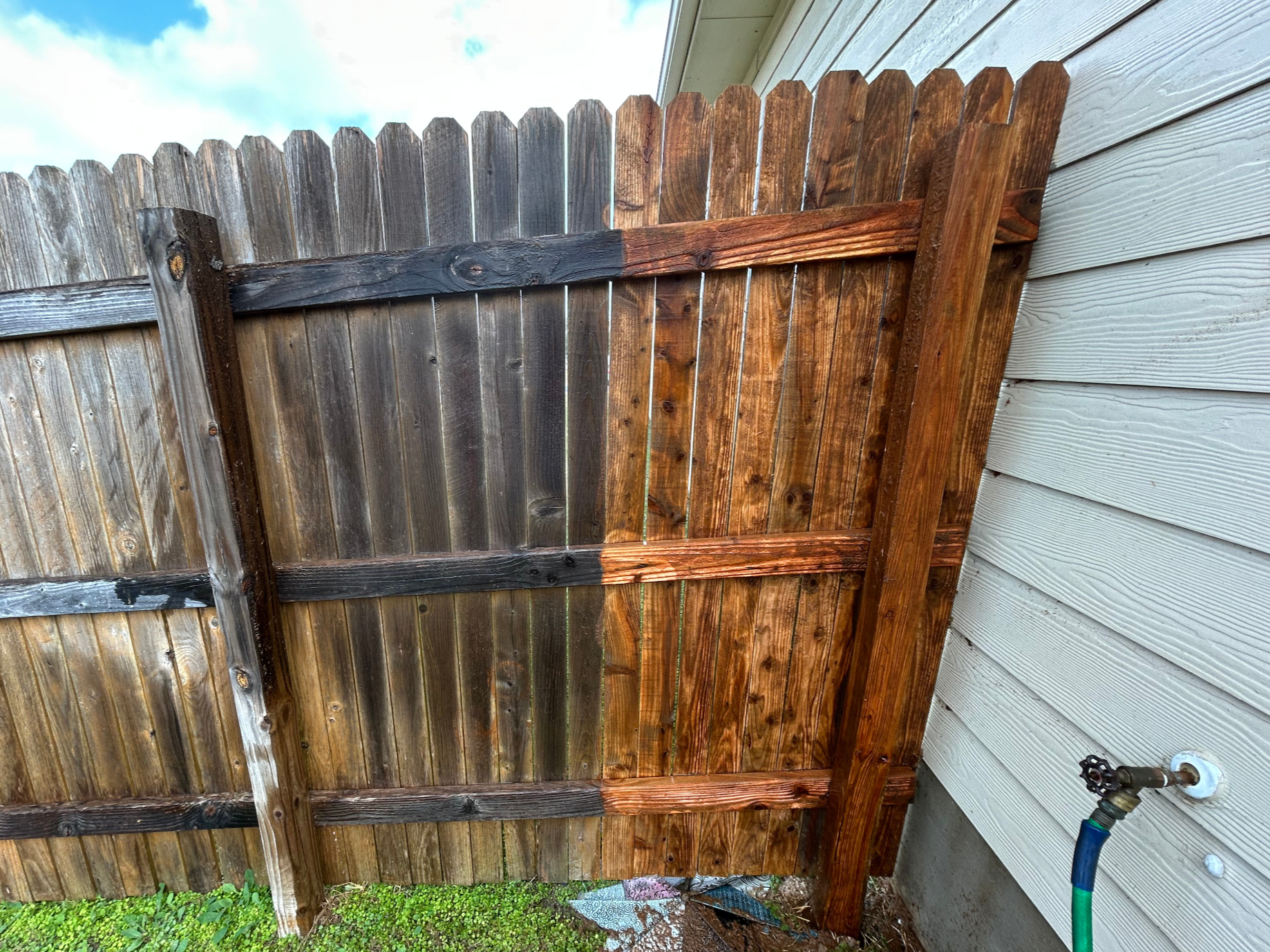 A wooden fence attached to the side of a house with white siding. The fence has a mix of new and older wood, with one section recently stained a darker color. There is a garden hose faucet on the house wall.