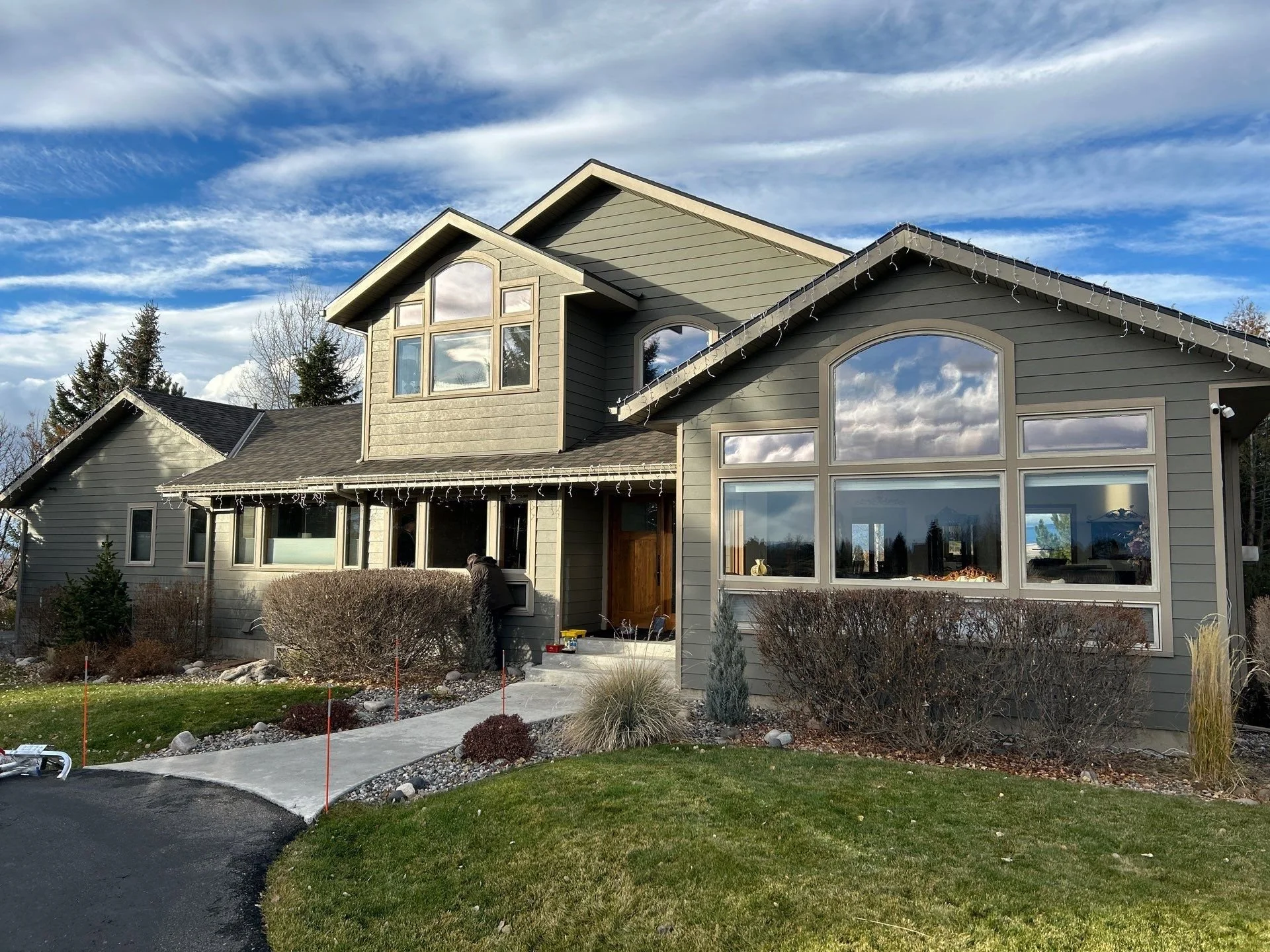 A large, modern house with a gray exterior, multiple windows, a front porch, and a landscaped lawn, with a partly cloudy sky above.