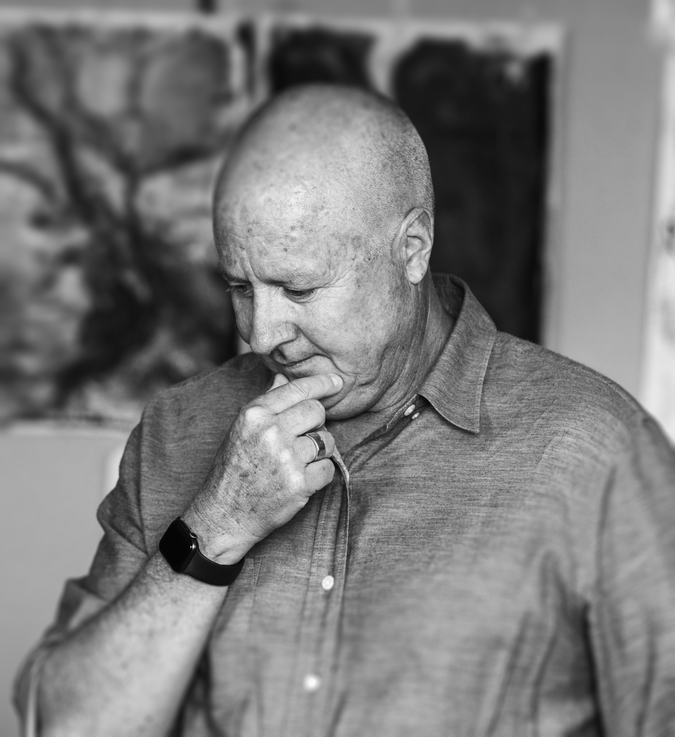 A man with a shaved head, wearing a gray shirt and a smartwatch, stands with hands clasped, looking down, in front of a blurred wall with three framed pictures.