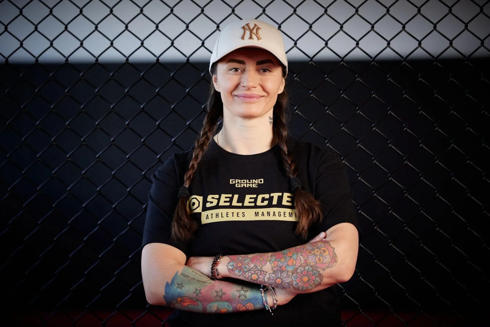 A woman with braided hair, tattoos on her arms, wearing a baseball cap and a black t-shirt, standing in front of a black chain-link fence and smiling.