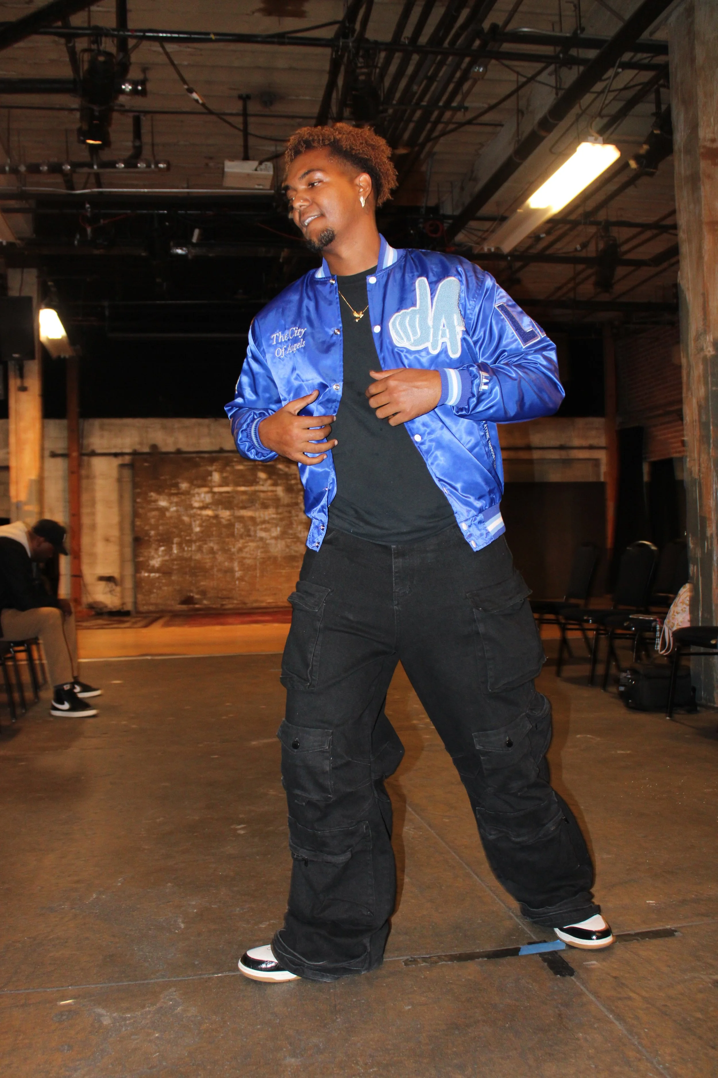 A young man with curly hair dancing in an industrial-style room with exposed ceiling pipes, wearing a bright blue satin baseball jacket, black cargo pants, and sneakers.