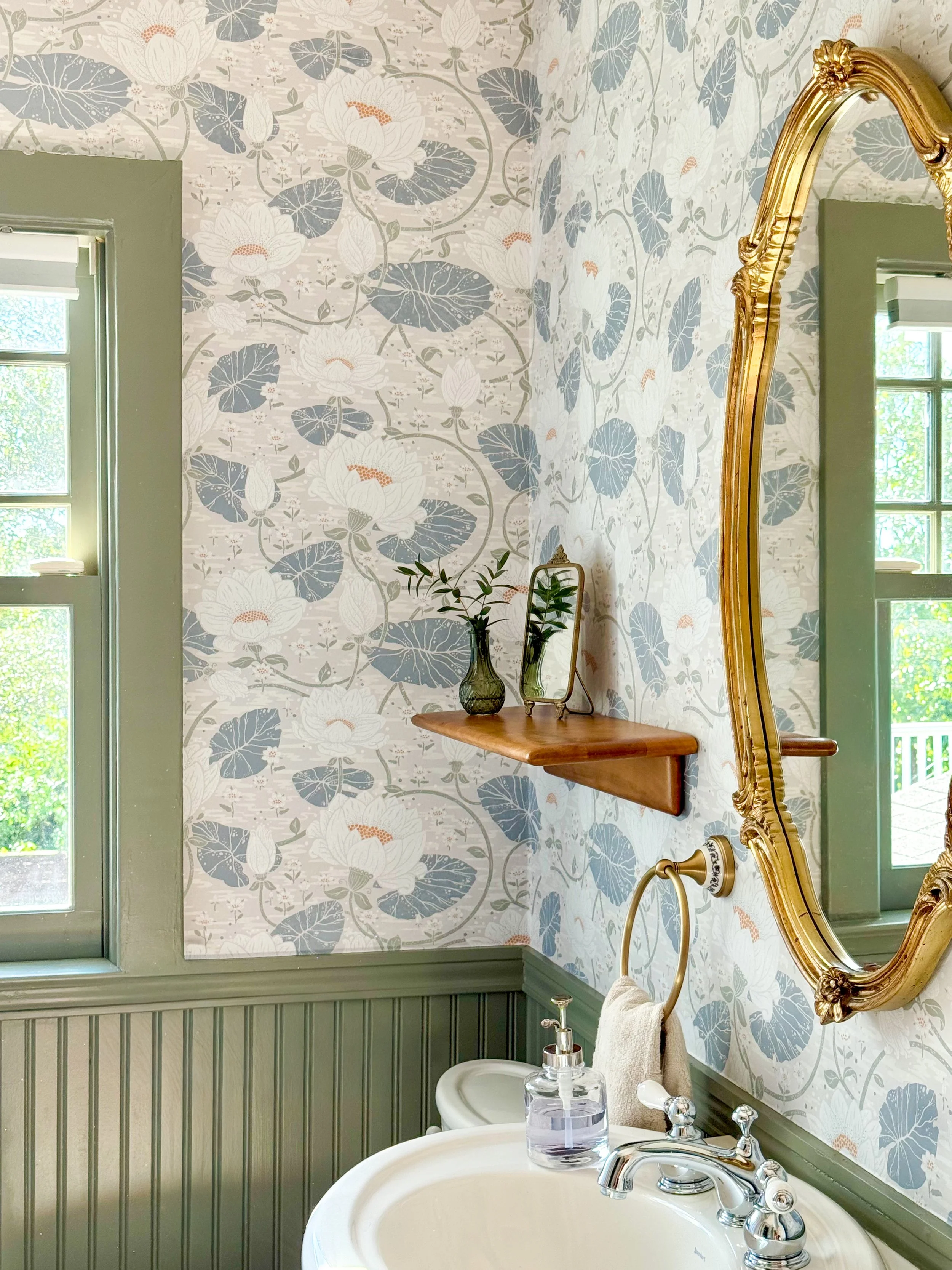 A bathroom with floral wallpaper featuring white and gray lilies, olive green beadboard wainscoting, and a vintage gold-framed mirror. A small wooden shelf holds two vases with green foliage, and a small mirror is also on the shelf. The bathroom has 