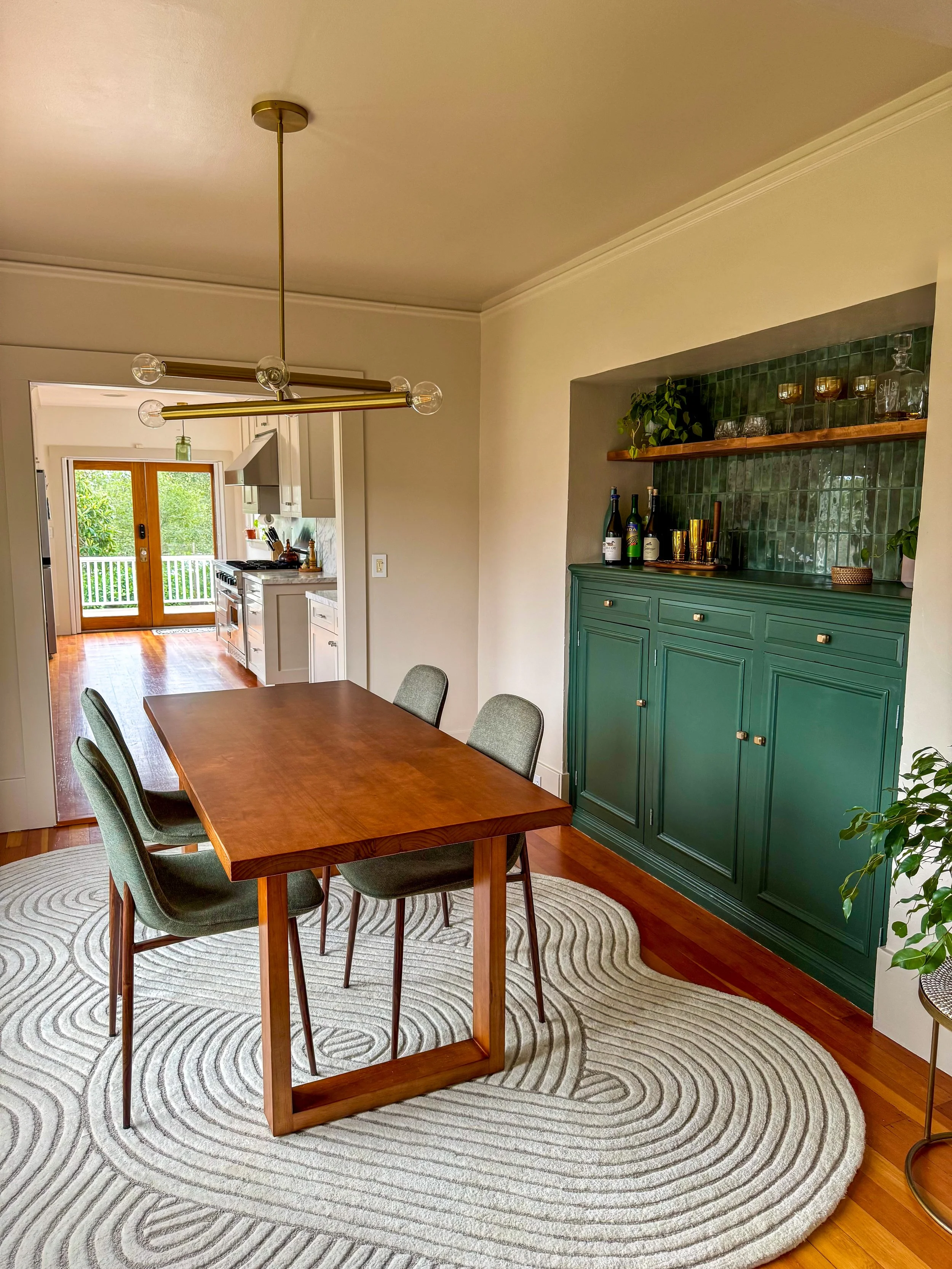 A wooden dining table with four green upholstered chairs in a dining room, featuring a green cabinet with decorative bottles and glasses, a green tiled wall, and a large round cream rug with abstract line patterns. In the background, there is a kitch