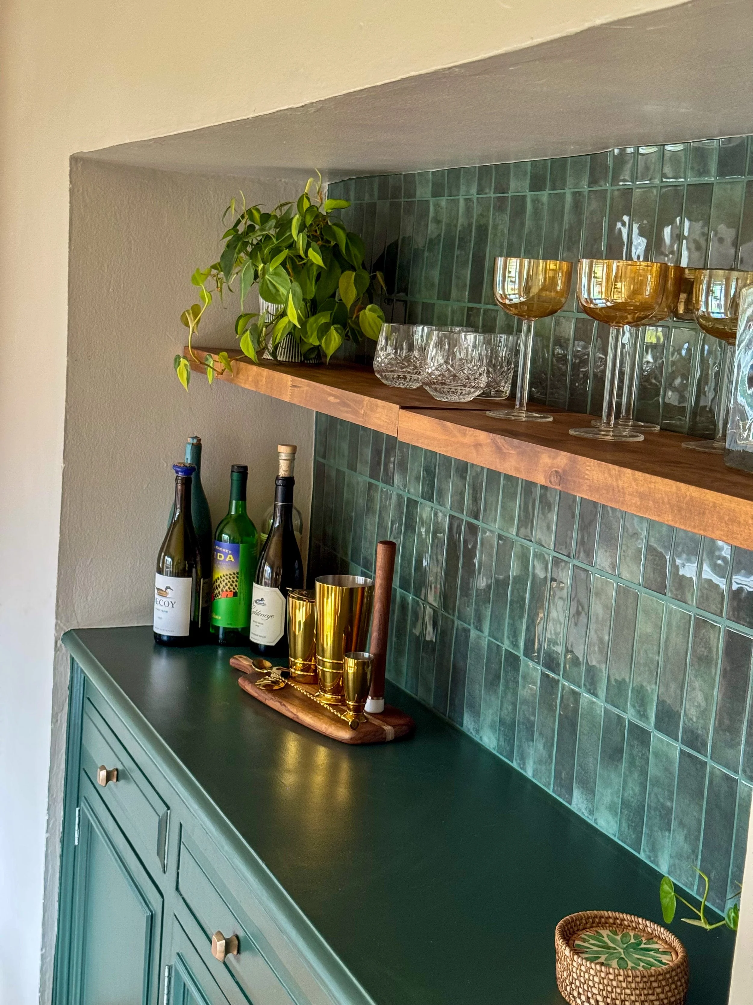 A green cabinet with gold knobs underneath a wooden shelf, decorated with bottles of alcohol, gold cups, and glassware, with green tiled wall and potted plant.