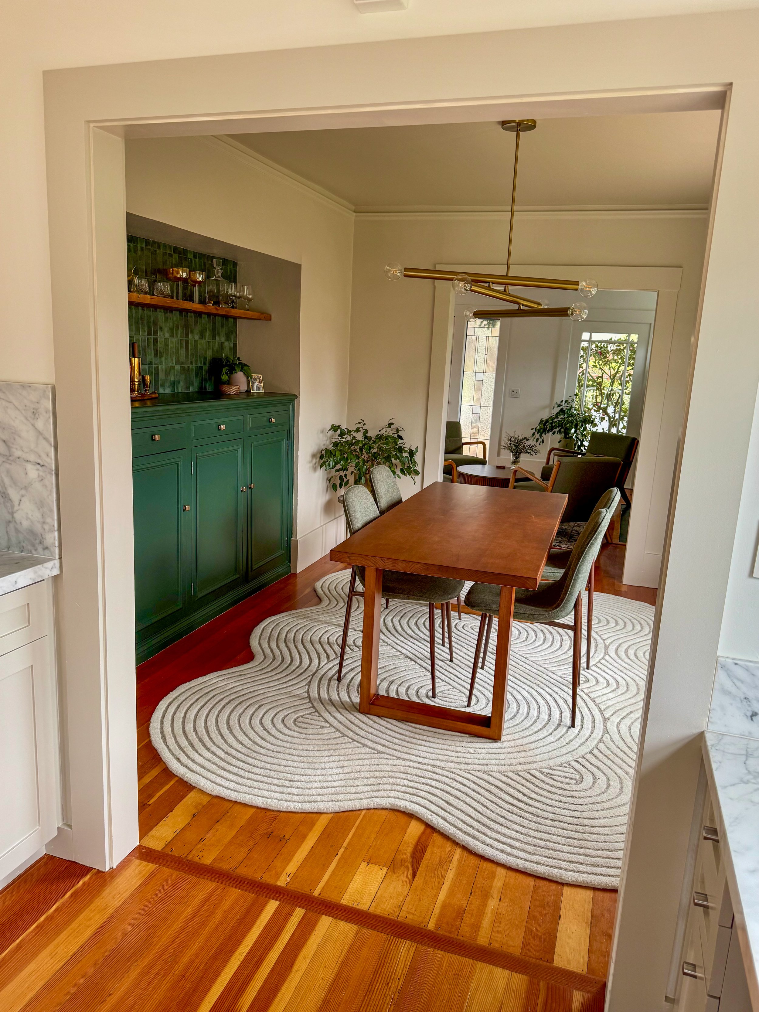A dining room with a wooden table and six upholstered chairs, a green cabinet with glassware, a gold modern chandelier, potted plants, and a cream and gray patterned rug on the hardwood floor.