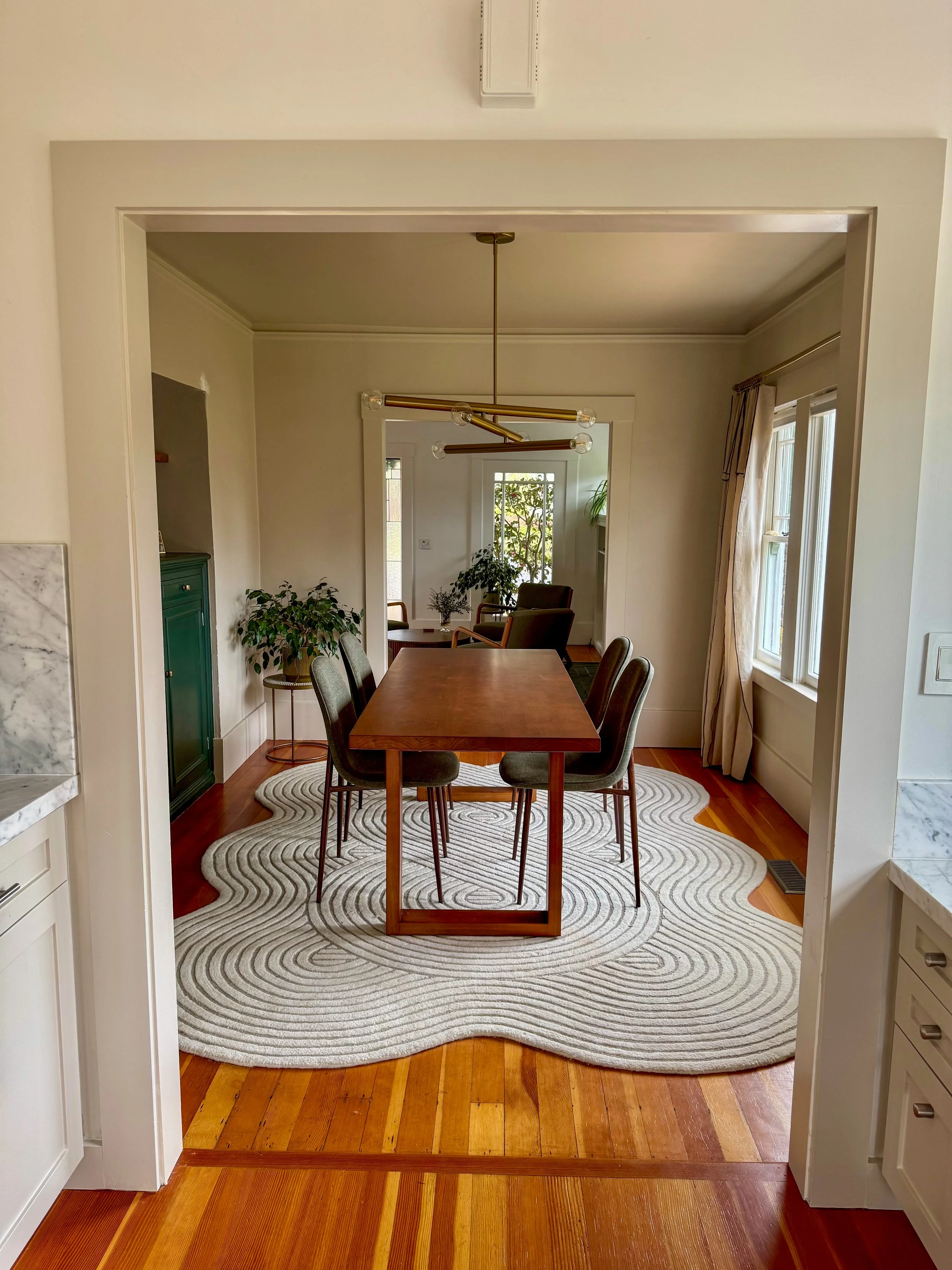 A dining room with a wooden table and six upholstered chairs on a circular, patterned beige rug. There are large windows with cream curtains and a modern gold chandelier hanging above the table. The room has wood flooring and some plants near the win
