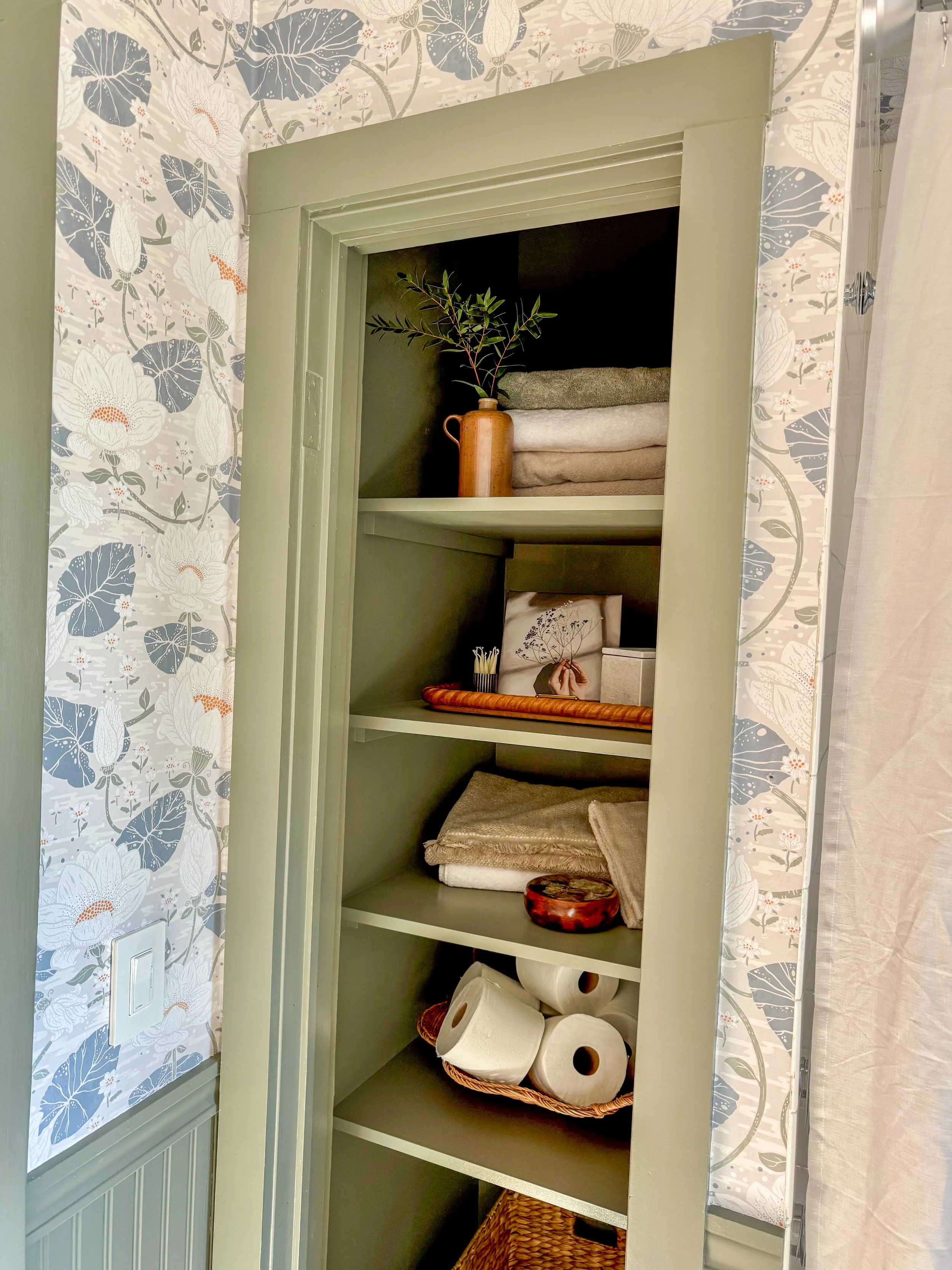 A green linen closet with four shelves containing folded towels, a potted plant, a framed picture, a decorative box, paper towels, and a woven basket at the bottom.