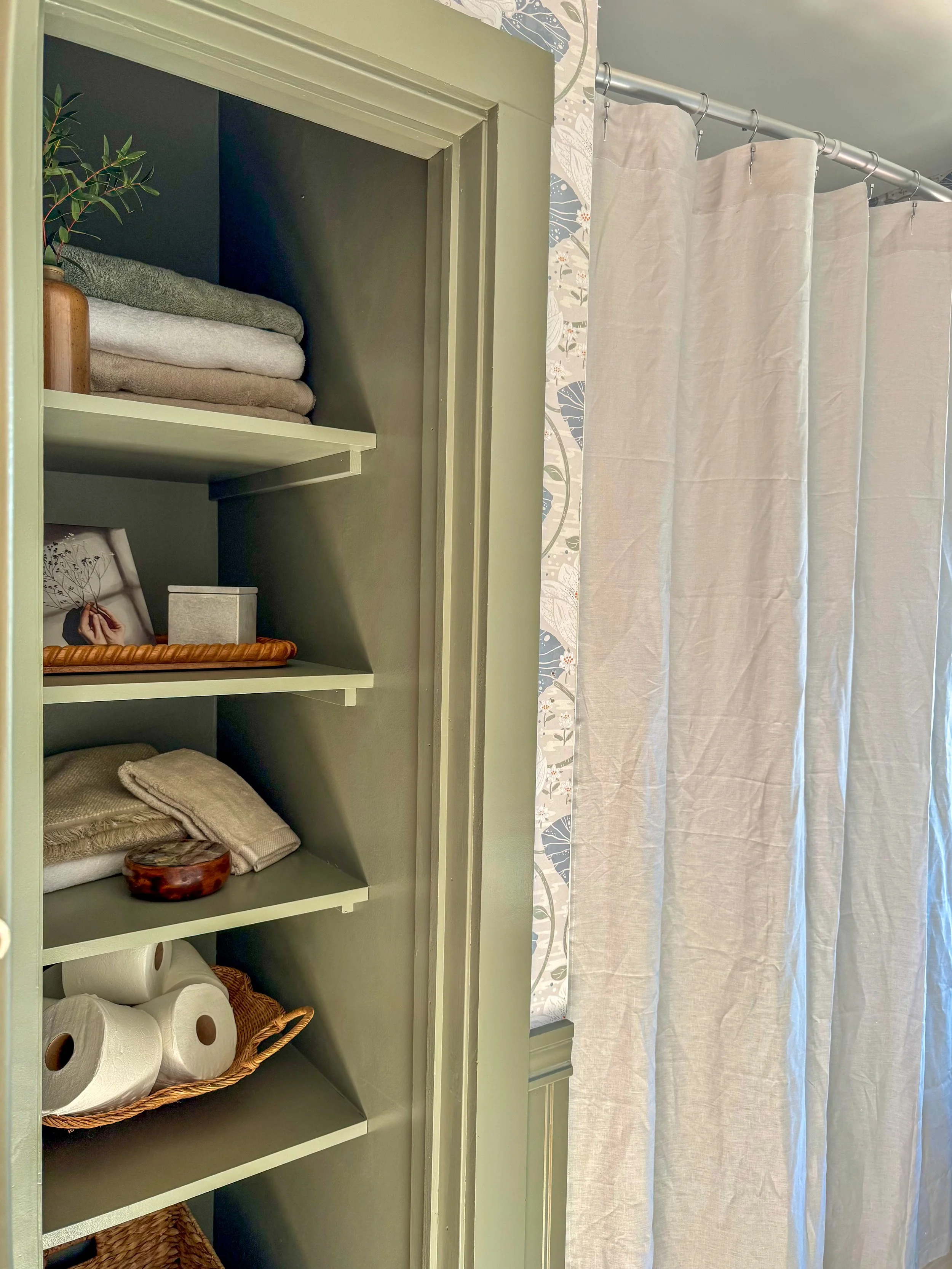 Bathroom shelves with folded towels, a framed picture, a small gray box, and toilet paper rolls in a bathroom with a white curtain and patterned wallpaper.