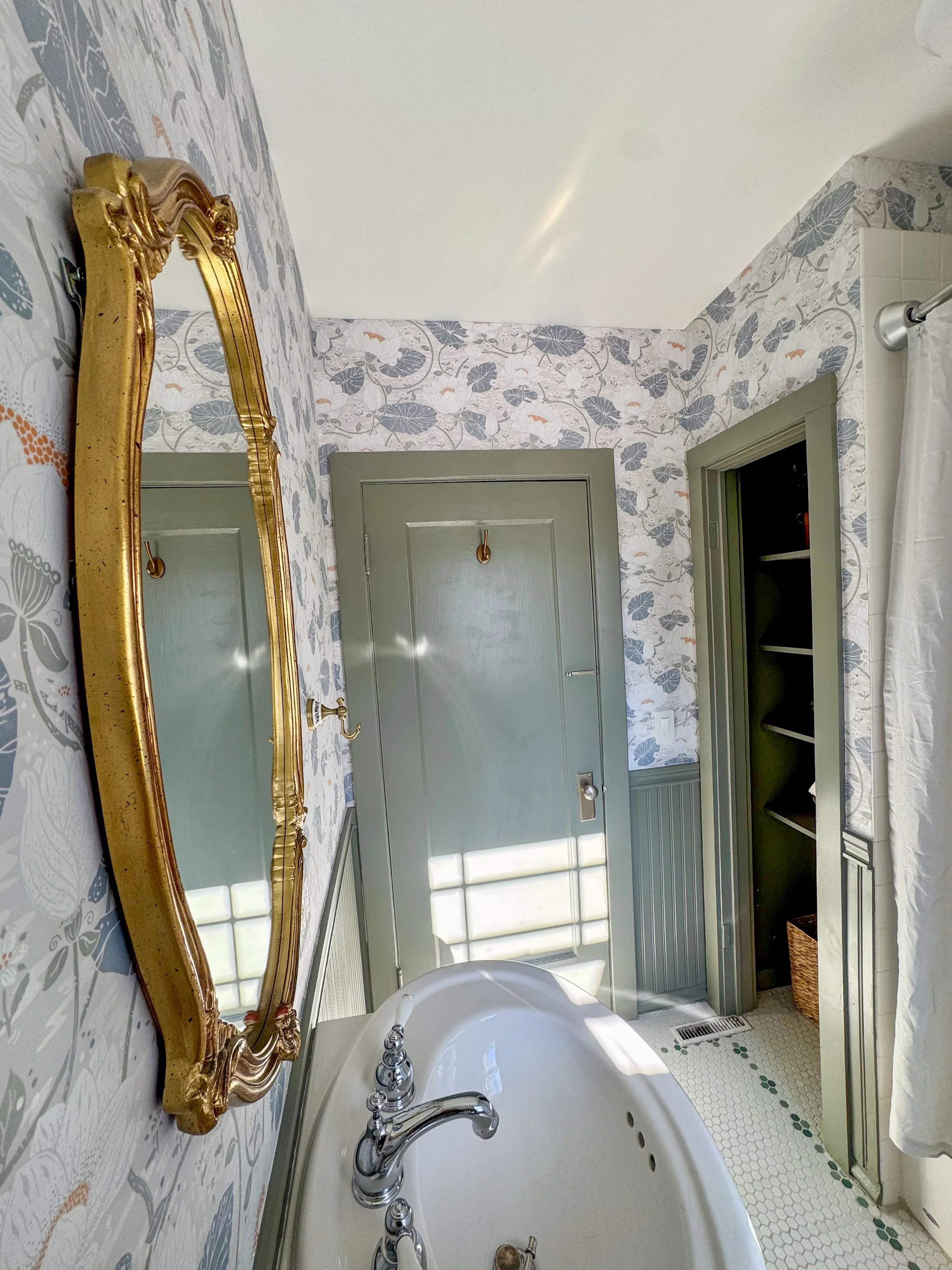 A vintage bathroom with a floral wallpaper, an ornate gold mirror, a bathtub with silver fixtures, and a green door and cabinet.