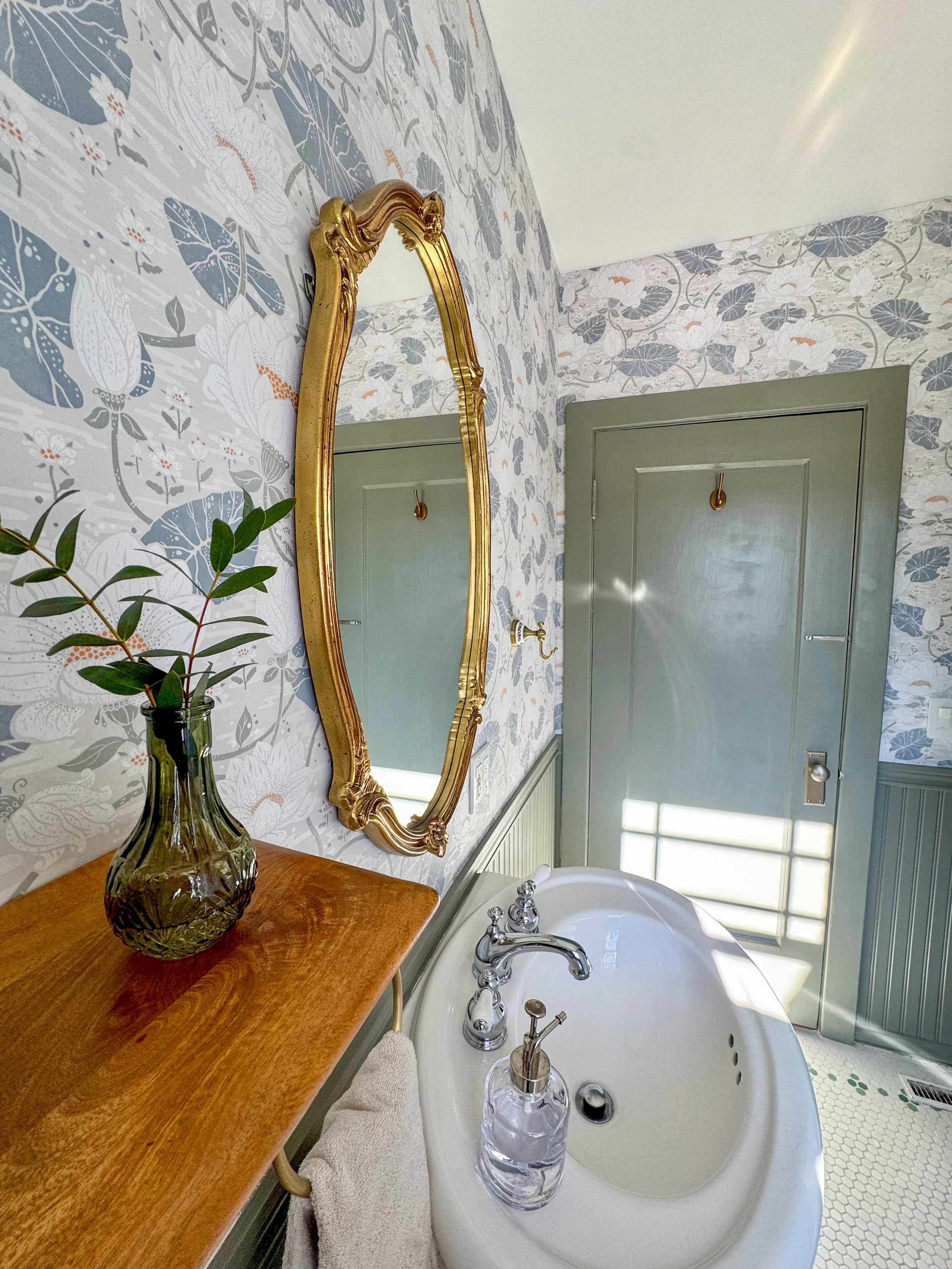 A vintage bathroom with floral wallpaper, a gold-framed mirror, a porcelain sink with chrome fixtures, a wooden shelf with a green vase containing green leaves, and a closed green door with a hook on it, illuminated by natural light.