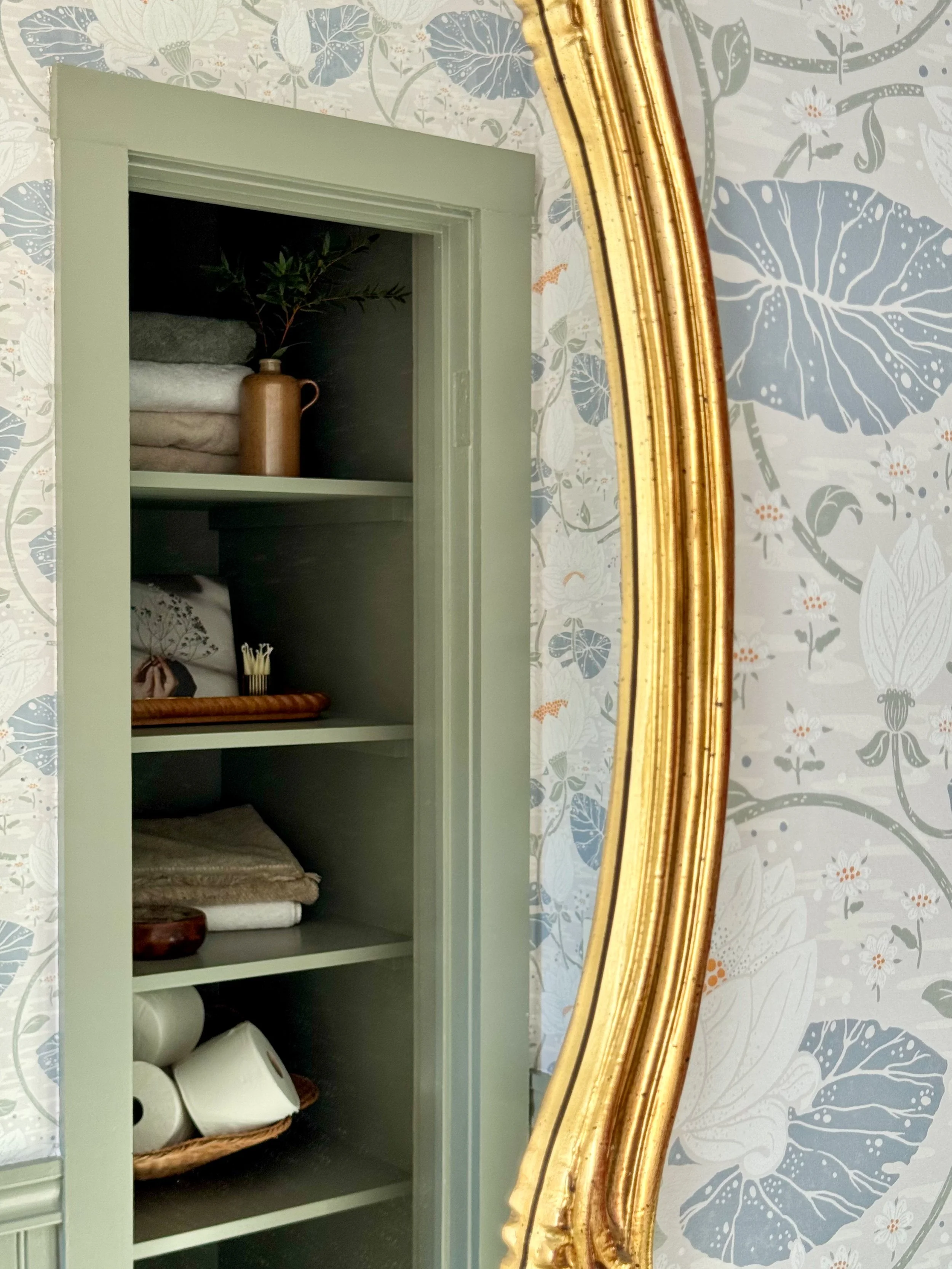 A reflection in a partial view of a mirror showing a green cabinet with shelves holding towels, a vase with a plant, a small bowl, and rolls of toilet paper, next to a wallpapered wall with a floral pattern.
