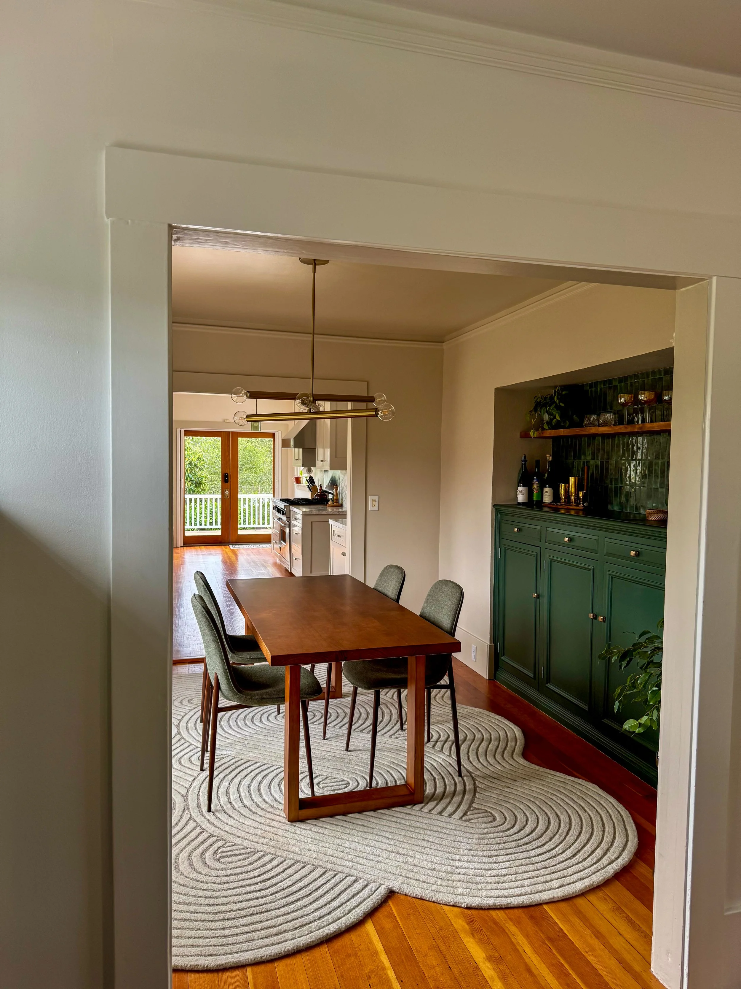A dining room with a wooden table, four green upholstered chairs, a patterned rug, a green sideboard, and a chandelier, with a view of a kitchen and balcony with greenery outside.