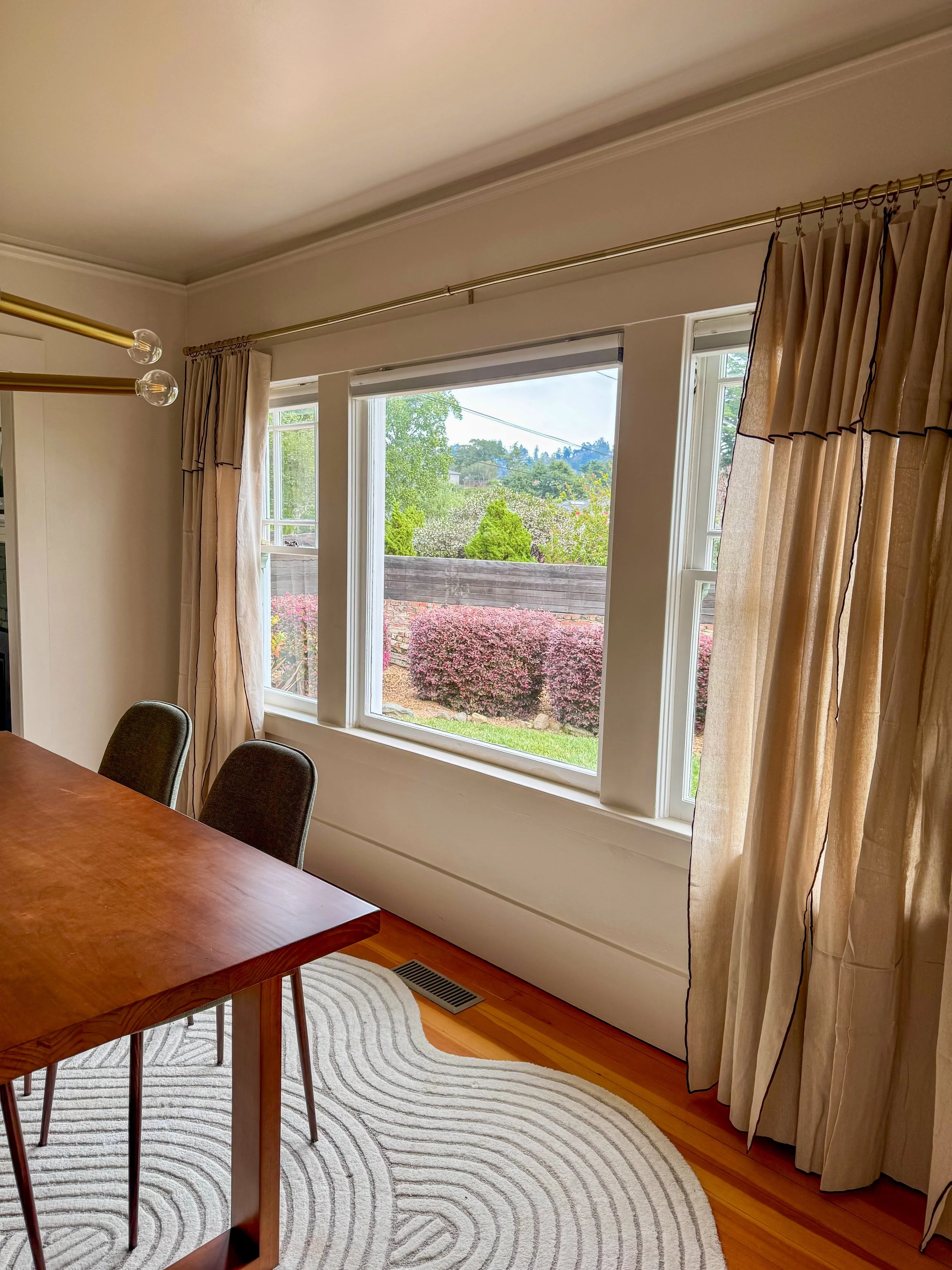 A dining area with a wooden table, dark upholstered chairs, a patterned white rug, large windows with beige curtains, and a view of bushes and trees outside.