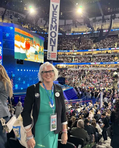 A woman in a turquoise dress and black blazer standing in a crowded large indoor event venue with a stage and large screen behind her displaying the word 'VERMONT' and the year '2024'.