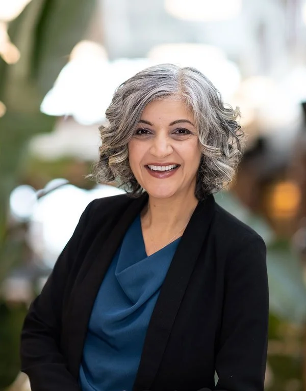 Smiling professional woman with curly gray hair wearing a black blazer over a blue blouse, standing indoors with a blurred background of natural light and greenery.