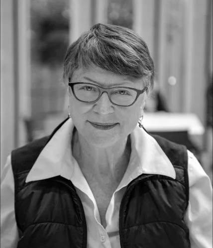 Black and white photo of a woman with short hair, glasses, wearing a white shirt and a black vest, smiling outdoors.