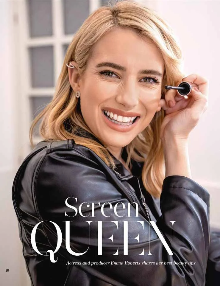 A woman with blonde hair smiling and applying makeup with a black eyeliner pen. She is wearing a black leather jacket.