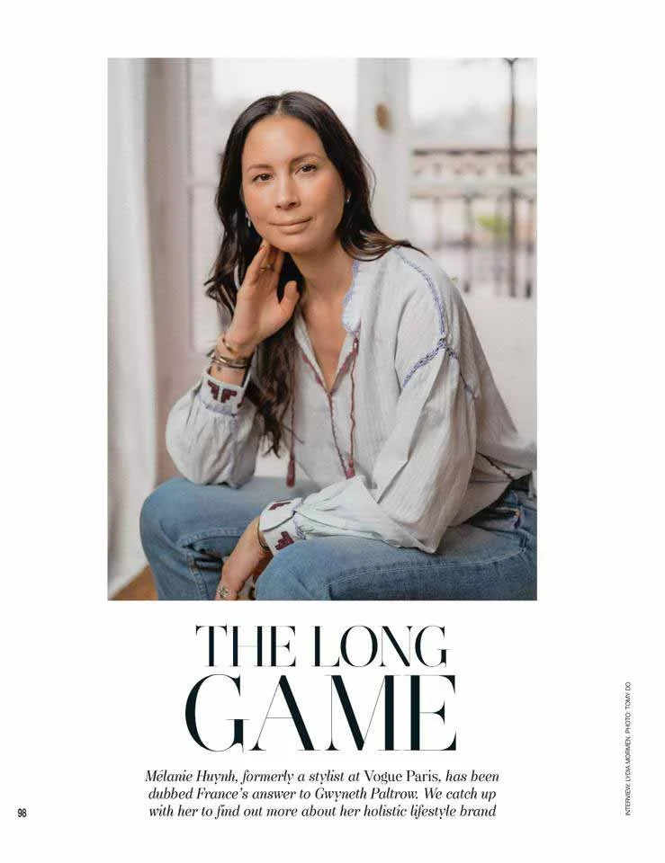 Portrait of a woman with long dark hair sitting on a chair indoors, wearing a light blouse and jeans, with a city view in the background.