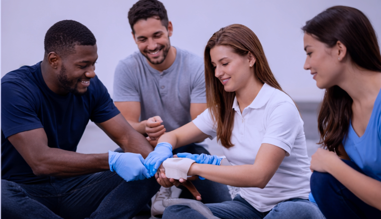 A group of four diverse people, including a woman with red hair and men with dark and light hair, participating in a first aid training session, practicing bandaging a person's arm with blue gloves.