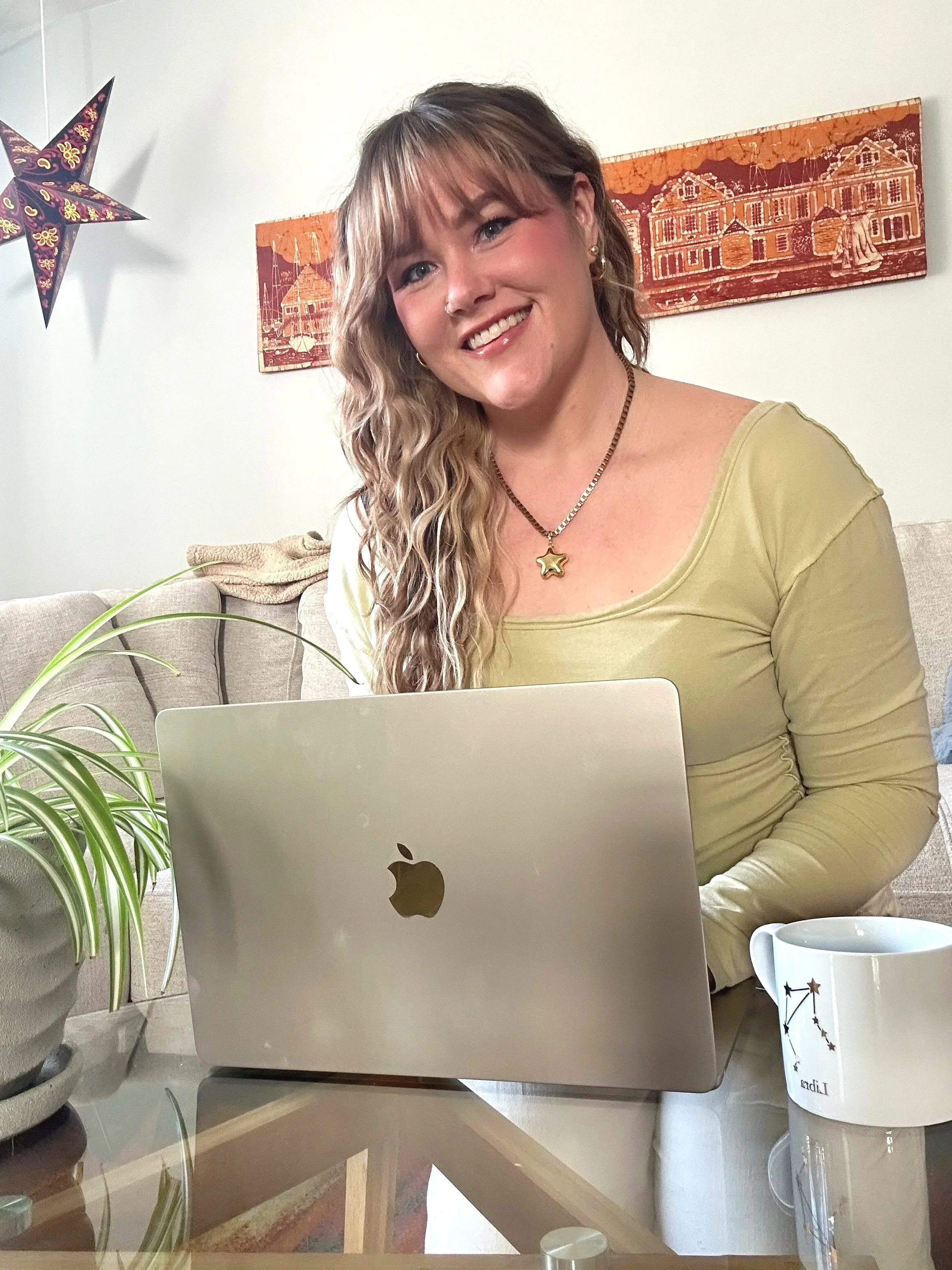A young woman with wavy blonde hair and a nose piercing smiling at the camera, sitting at a table with a silver MacBook, a green potted plant, and a white mug with constellation design, in a cozy home setting with wall art and a star-shaped decoration in the background.