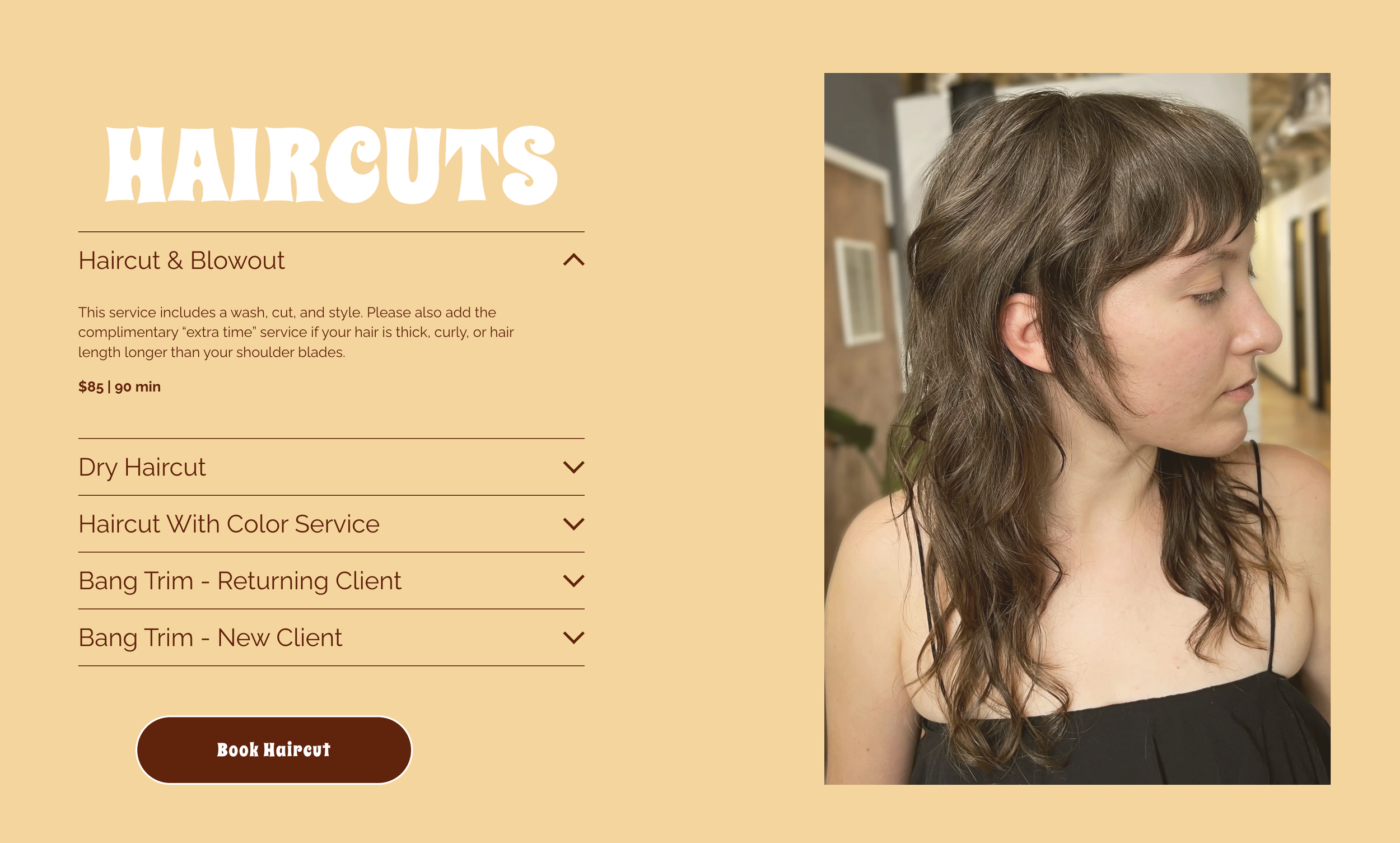 A woman with curly brown hair wearing a black sleeveless top, sitting in a salon with hairdressing equipment in the background.
