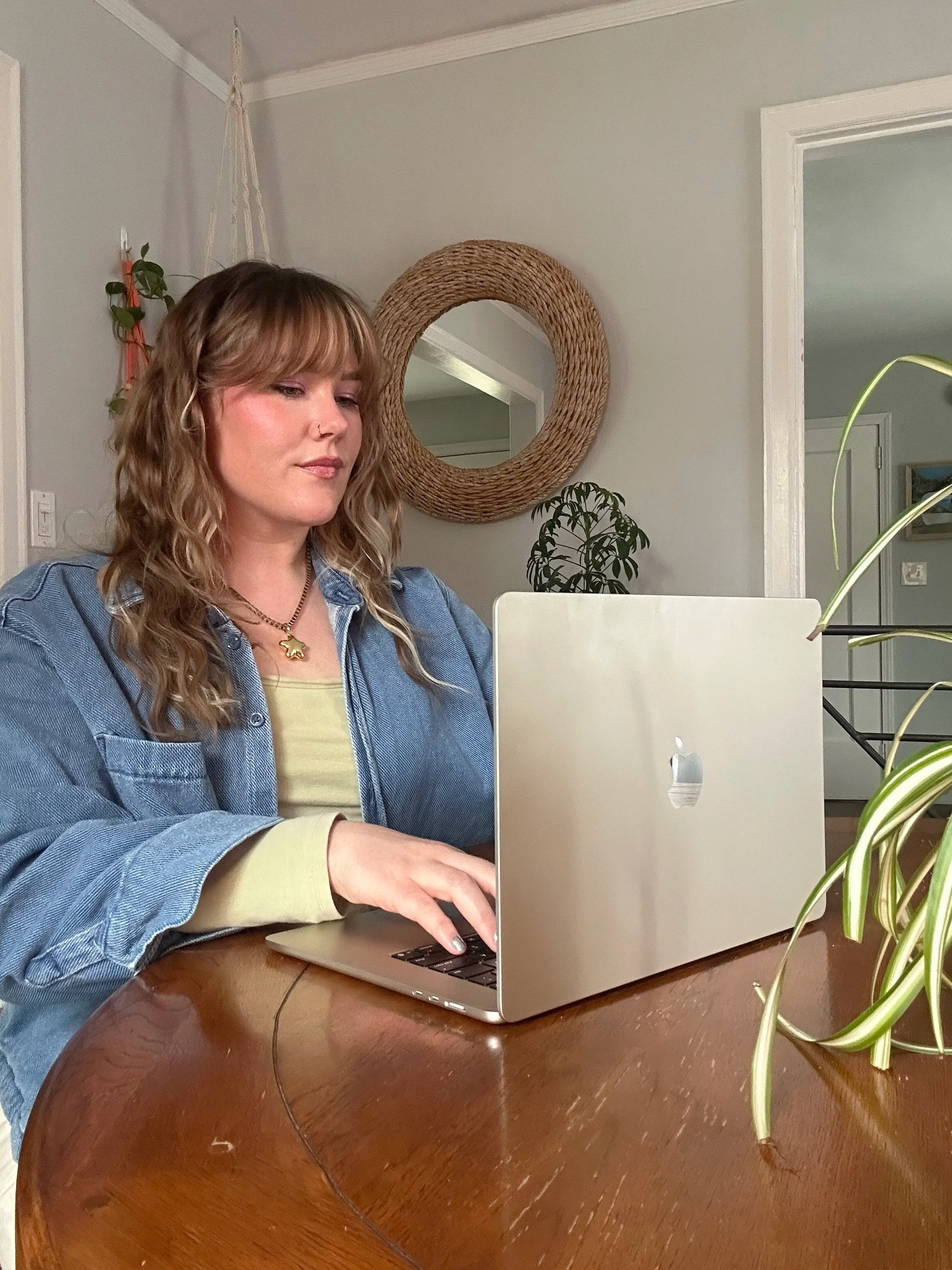 A woman with curly blonde hair working on a silver MacBook laptop at a wooden table in a cozy, well-decorated room with a round woven wall mirror, plants, and framed artwork in the background.