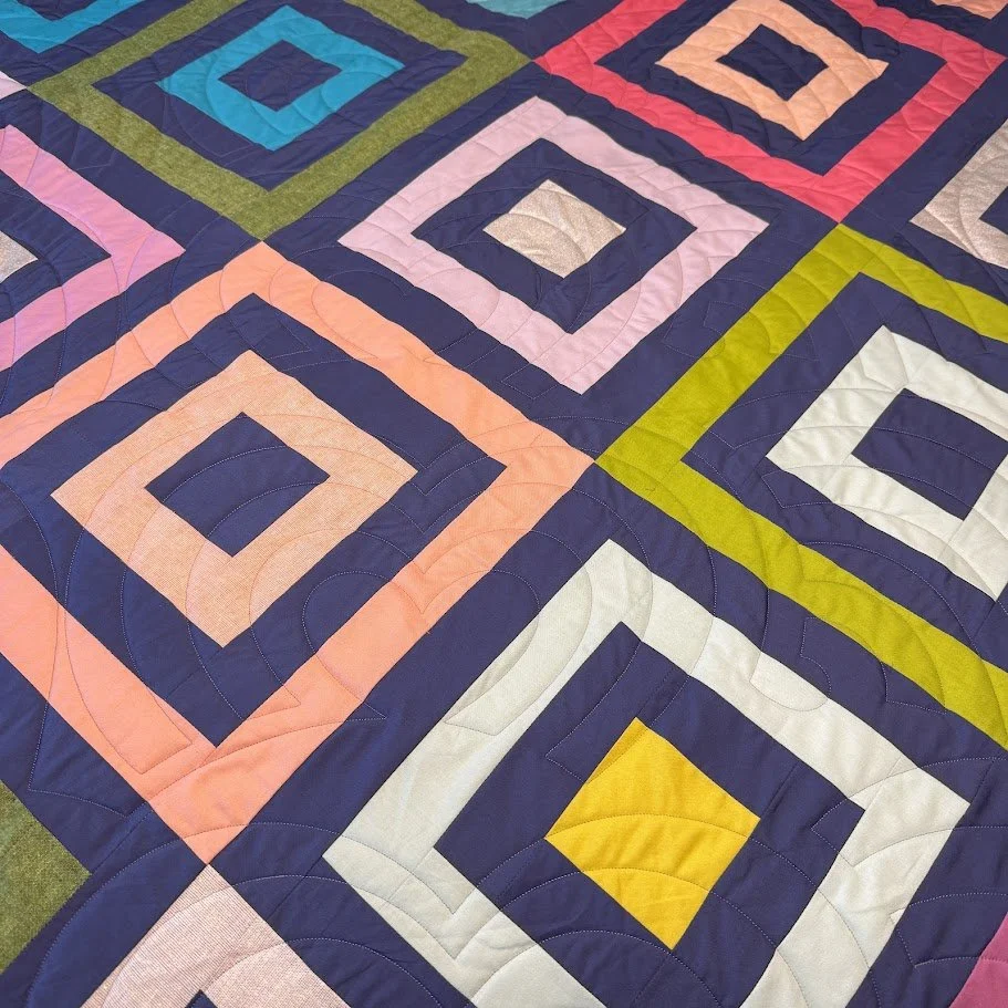 Close-up of a colorful patchwork quilt made of square blocks with concentric diamond patterns in various colors including pink, blue, yellow, green, and white.