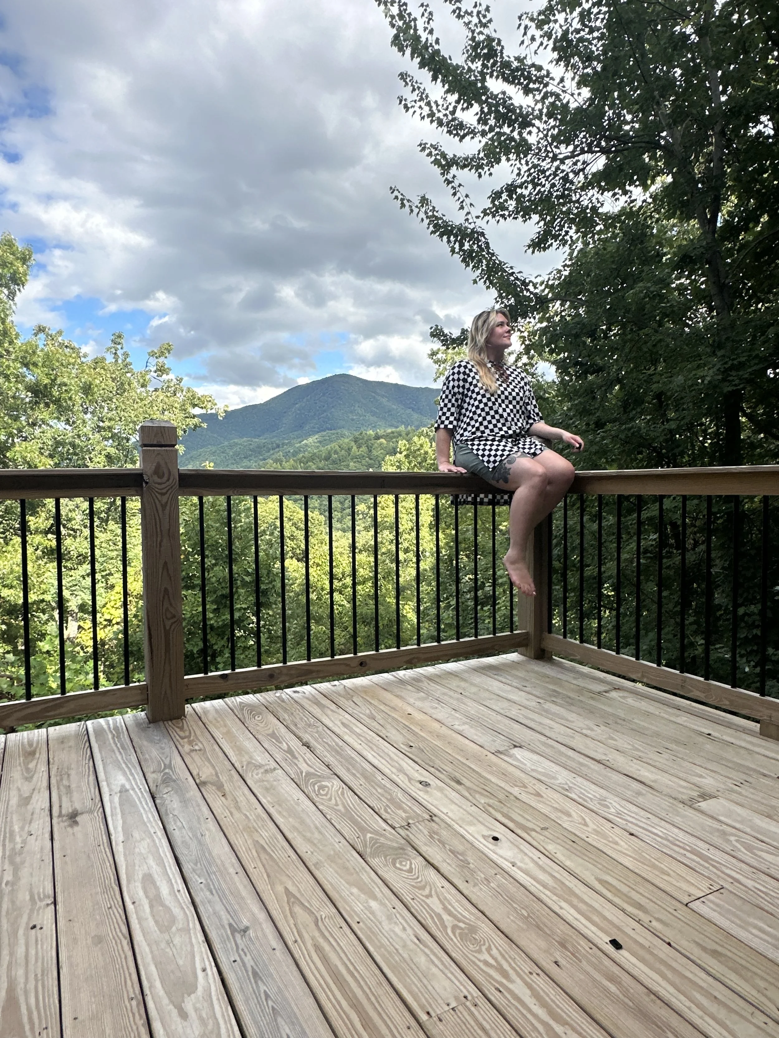 A woman sitting on a wooden deck railing with a scenic mountain and forest background, wearing a black and white checkered shirt and shorts.