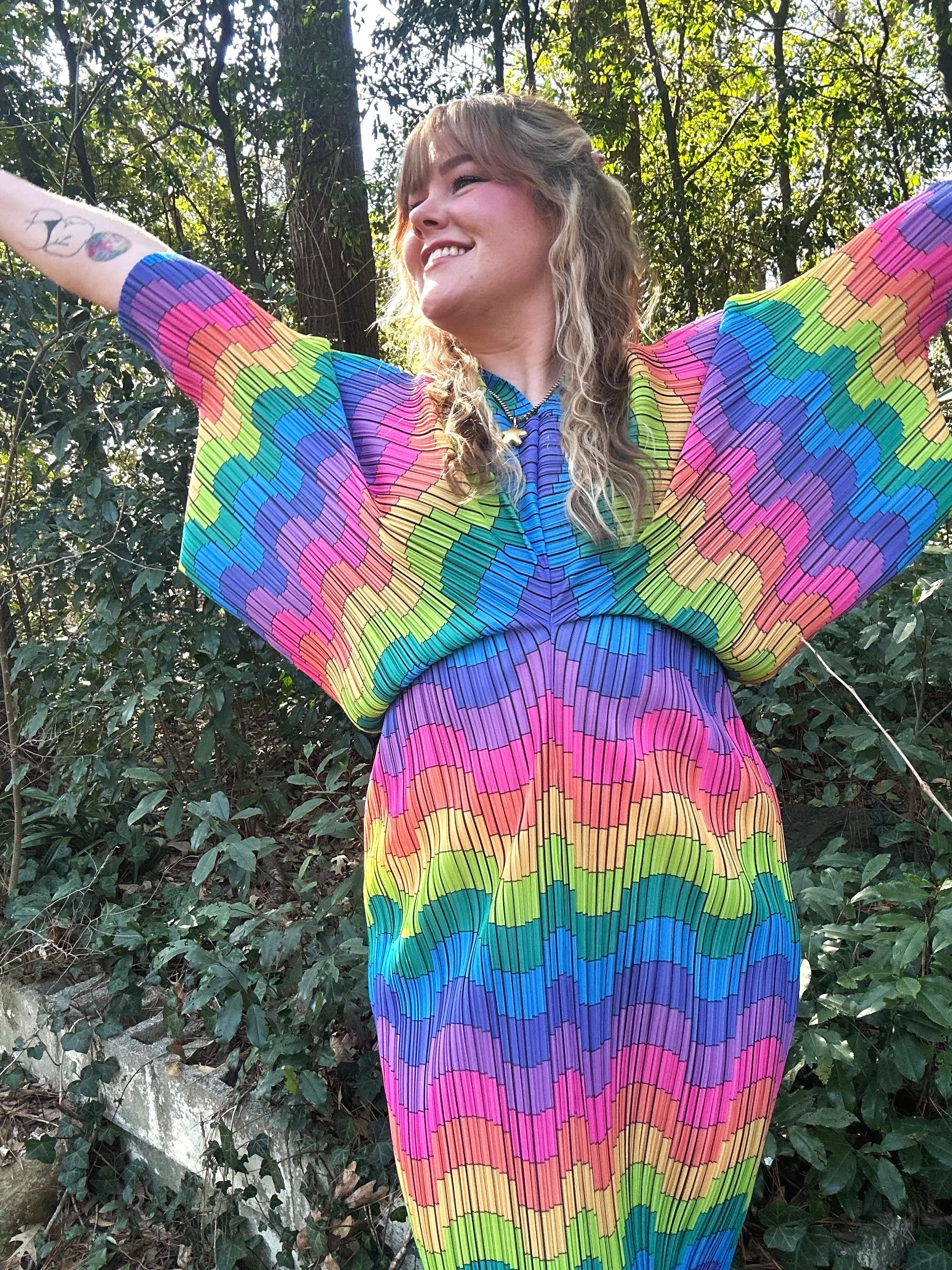 A smiling woman wearing a colorful, rainbow-patterned dress with her arms raised in front of a wooded outdoor background.