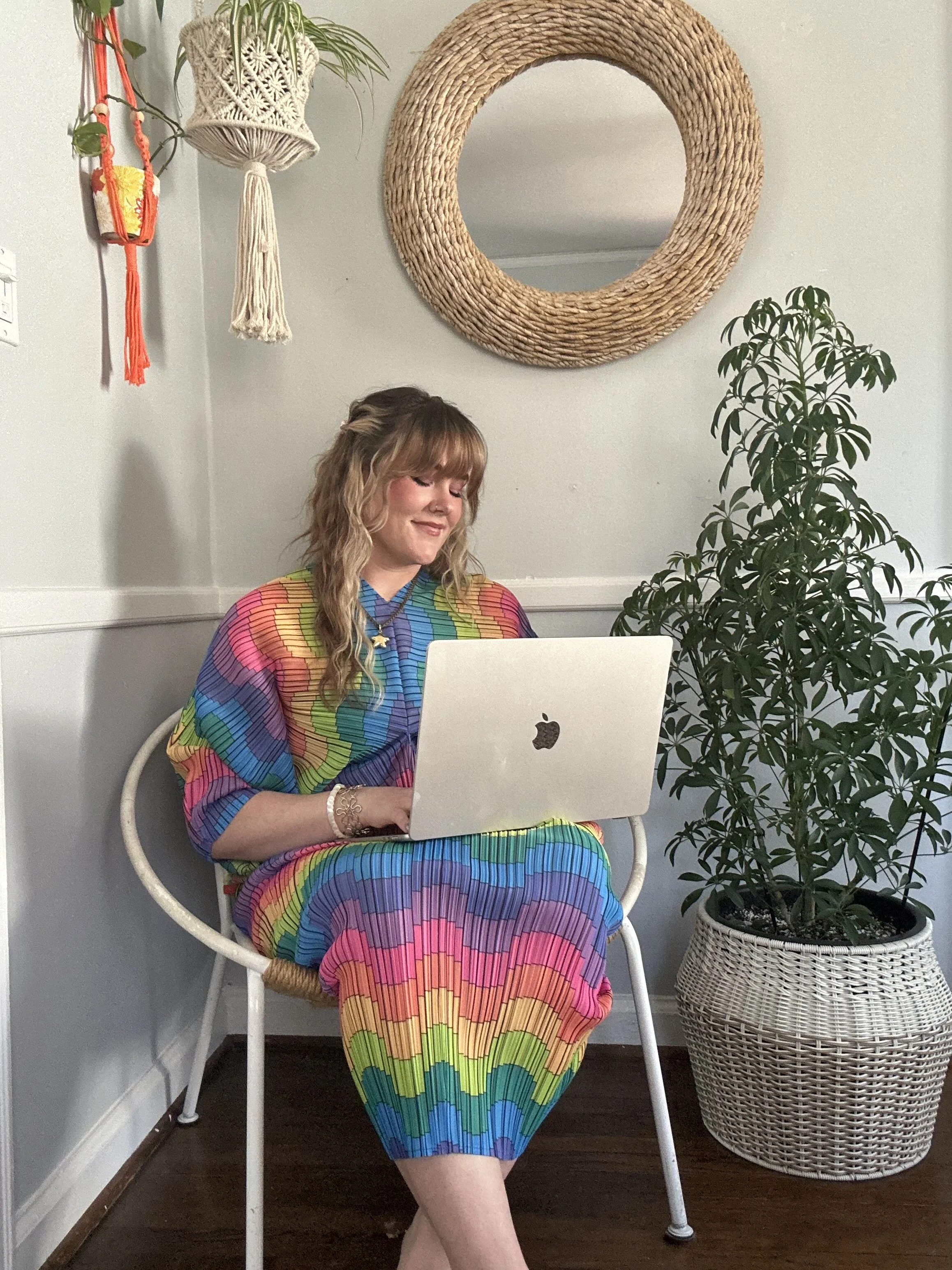 A woman sitting in a white chair with a rainbow-colored dress, working on a silver MacBook laptop. She is in a room with a large plant in a white woven basket, a wicker mirror on the wall, and hanging decor including a macrame plant holder and a small wall hanging.