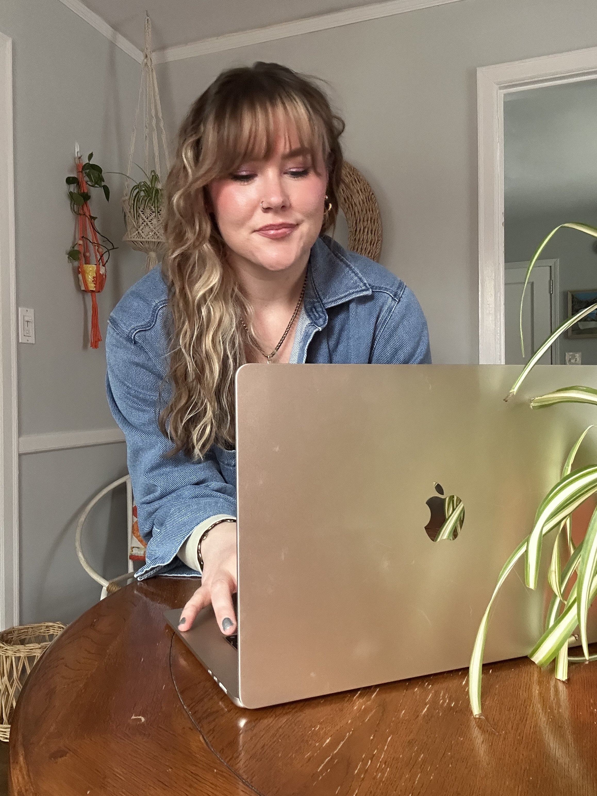 A woman with wavy blonde hair and a nose ring sits at a wooden table using a silver MacBook laptop, wearing a denim jacket.