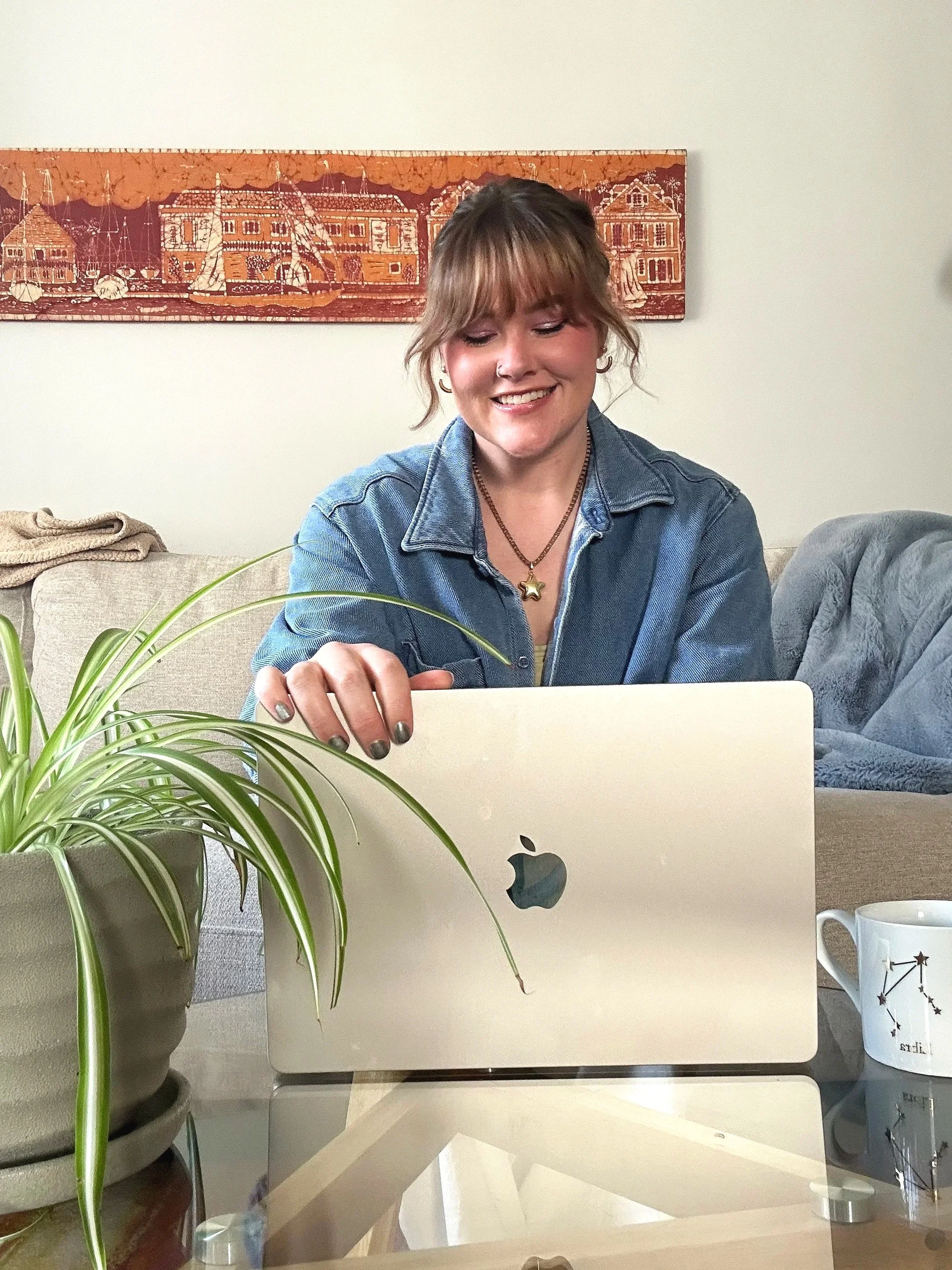 A woman is sitting at a glass table with a silver MacBook in front of her, smiling and touching the laptop. There is a potted plant, a coffee mug with constellation design, and a couch with a blanket on it in the background, along with a red and white painting on the wall.