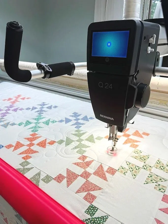 A Bernina quilting machine with an electronic screen, sewing colorful geometric quilt blocks on a white fabric.