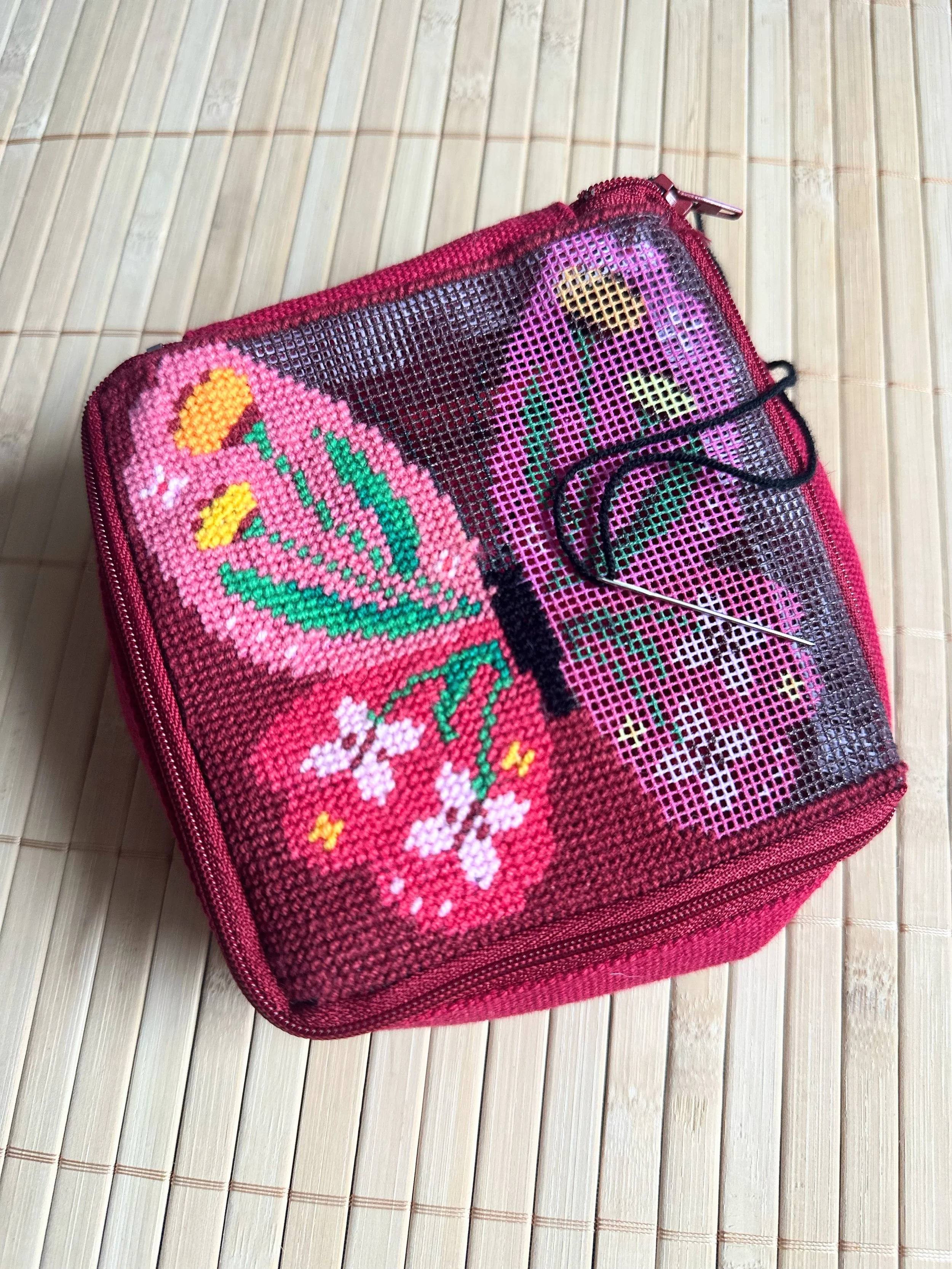 A pink zippered pouch with a pixel art butterfly and flower design under a mesh covering, placed on a bamboo mat.
