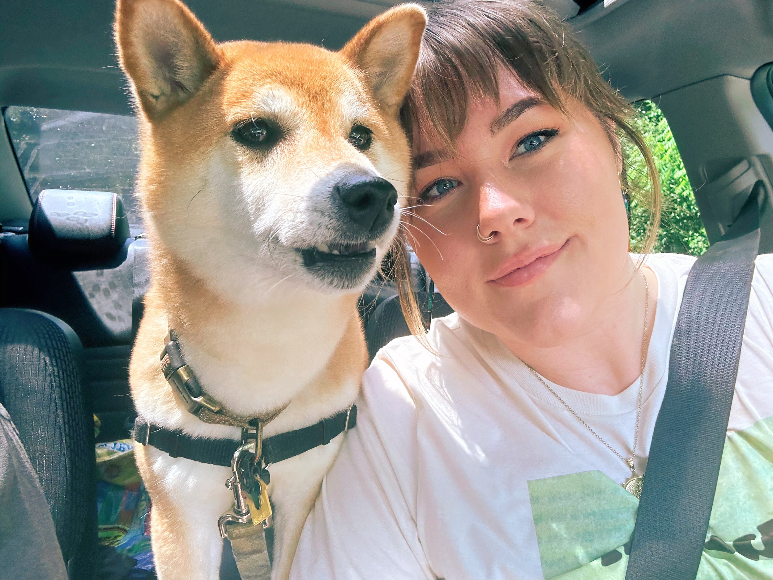 A woman smiling and a Shiba Inu dog sitting in a car, both looking at the camera.