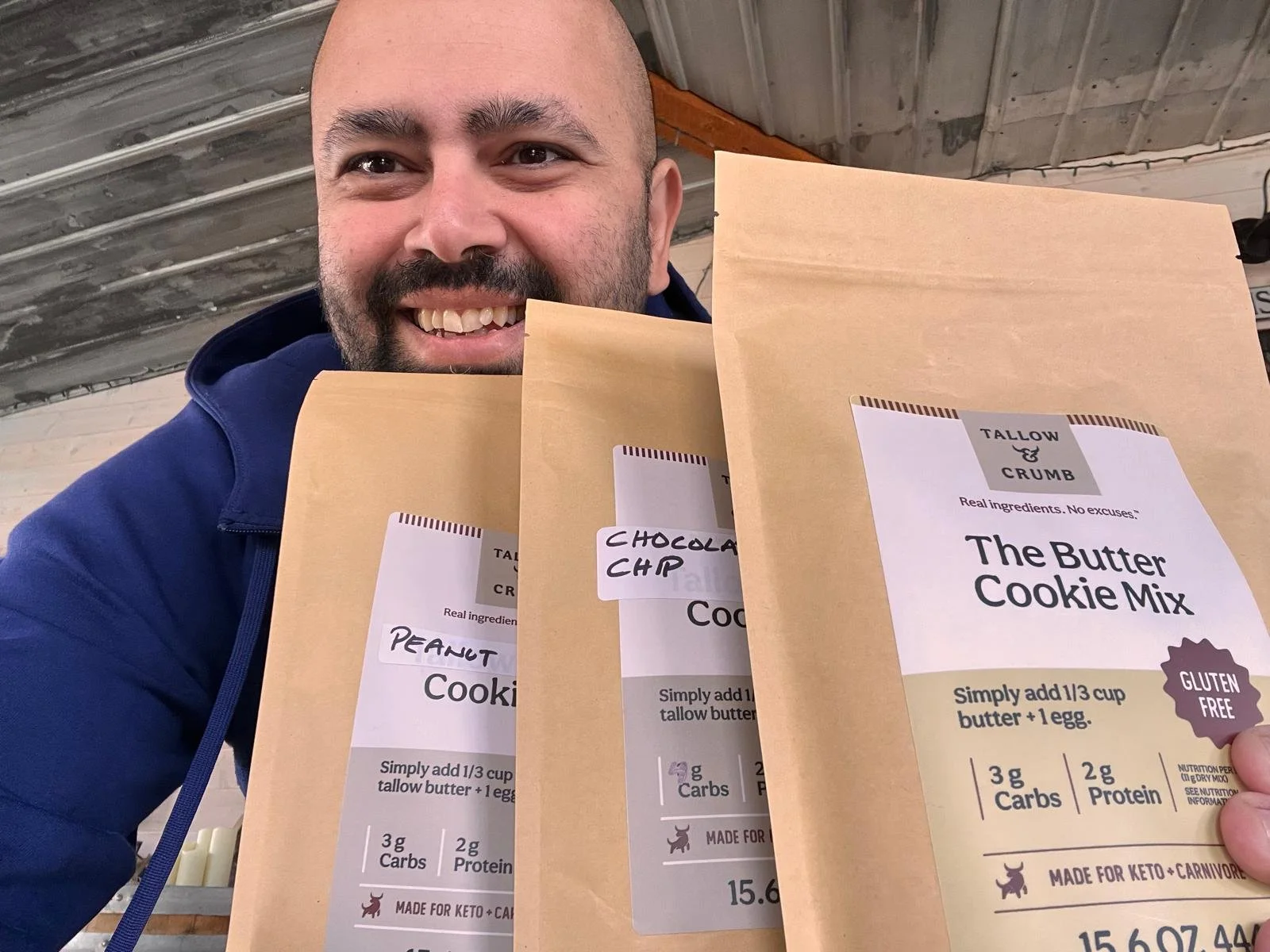 A man smiling while holding three different cookie mix packets. The visible packet labels read 'Tallow & Crumb,' 'Chocolate Chip,' and 'Peanut.' The packets have a beige/brown color with nutritional information and ingredients listed. The man is wearing a blue jacket and is standing indoors with a metal roof ceiling in the background.