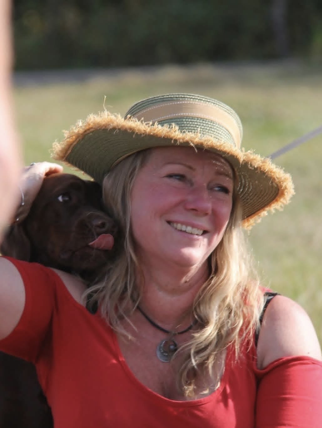 A woman wearing a large brim sun hat and a red top, smiling and holding a chocolate Labrador puppy.