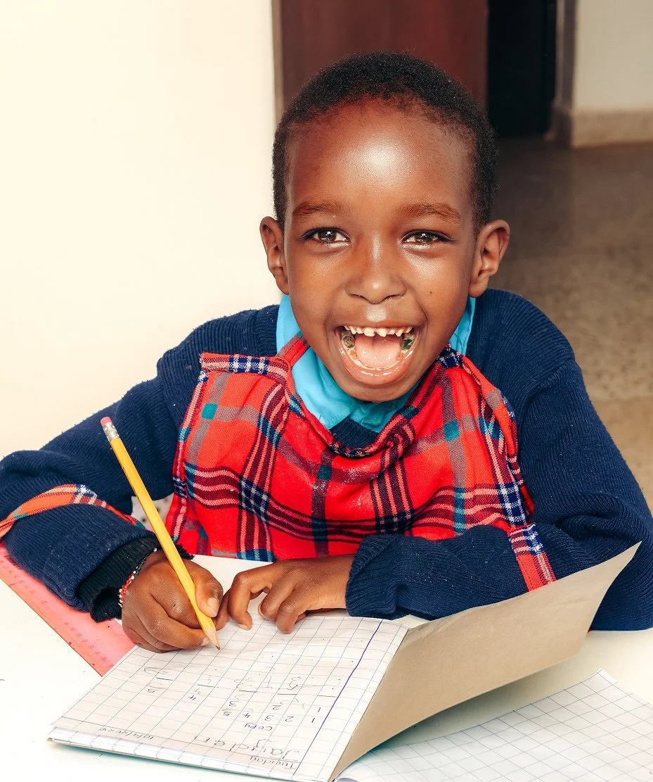 A young boy with a big smile, wearing a blue sweater and a red plaid scarf, sitting at a desk with a pencil in his hand and a notebook in front of him, appearing happy and engaged in a learning activity.