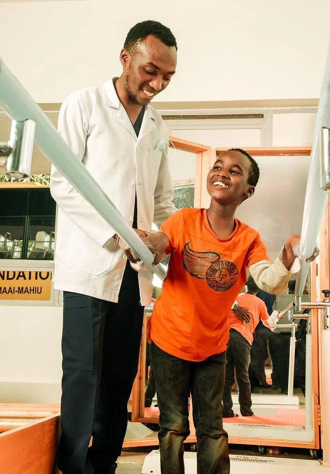 A young boy with a big smile holding onto parallel bars, assisted by a man in a white coat, likely a healthcare professional, in a rehabilitation or medical facility.