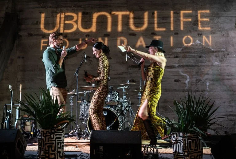 Three performers on stage at a live event, with a drum set in the background and the words 'UBUNTULIFE FESTIVAL' projected on the wall behind them. The two women are dressed in animal print dresses, one with a wide-brimmed hat, while the man is dress