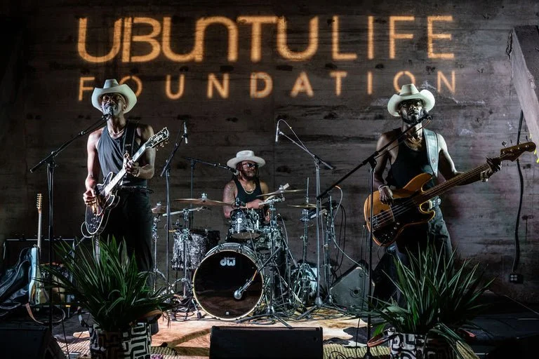 Three musicians performing on stage with a logo 'Ubuntu Life Foundation' projected on the wooden wall behind them, each wearing cowboy hats and playing instruments.
