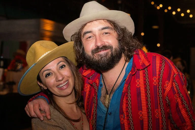 Two people smiling and hugging at a festive gathering, wearing colorful shirts and wide-brimmed hats in an indoor setting with string lights.